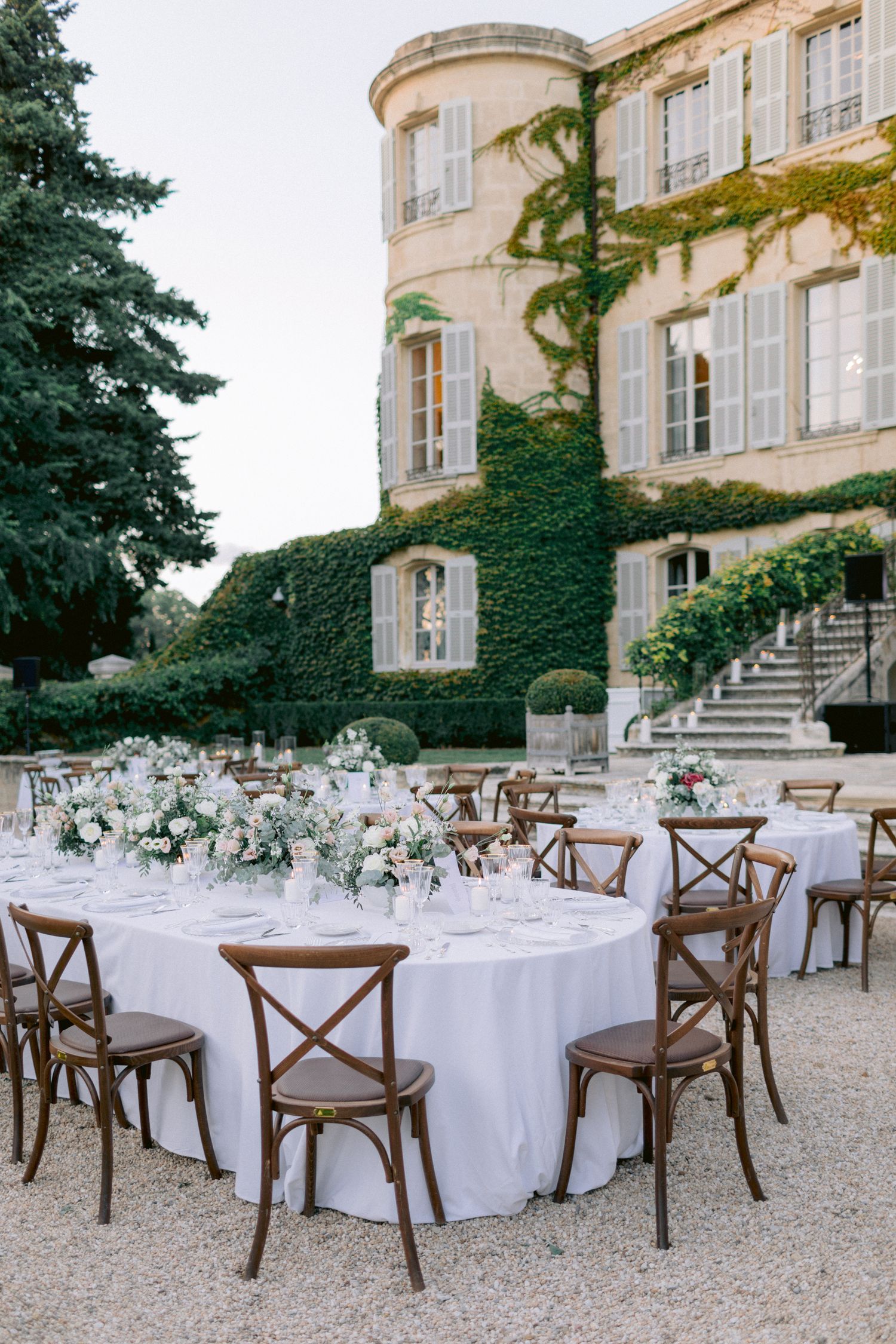 Elegant outdoor wedding reception setup with white linens and cross-back chairs at a French chateau.