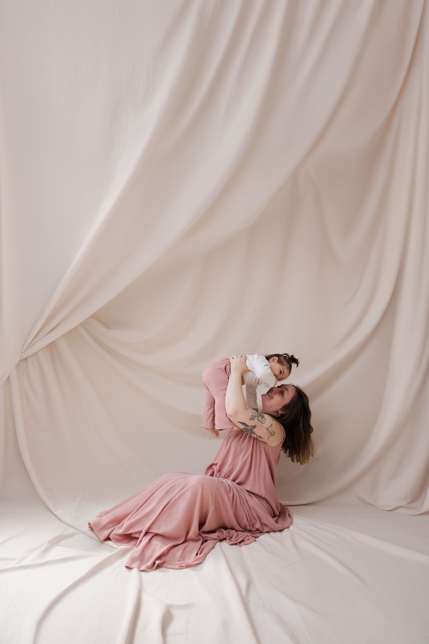 A woman in a pink dress sits on the floor lifting a baby against a flowing cream fabric backdrop.