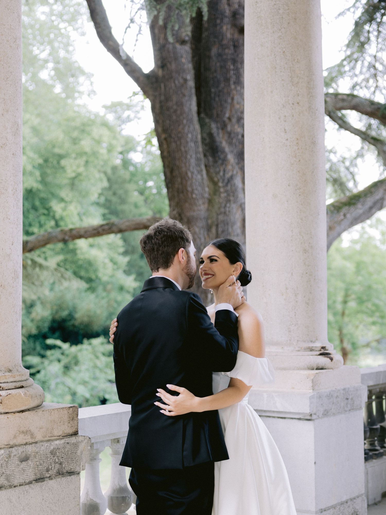 A romantic moment between a couple in formal attire standing between elegant white columns outdoors.
