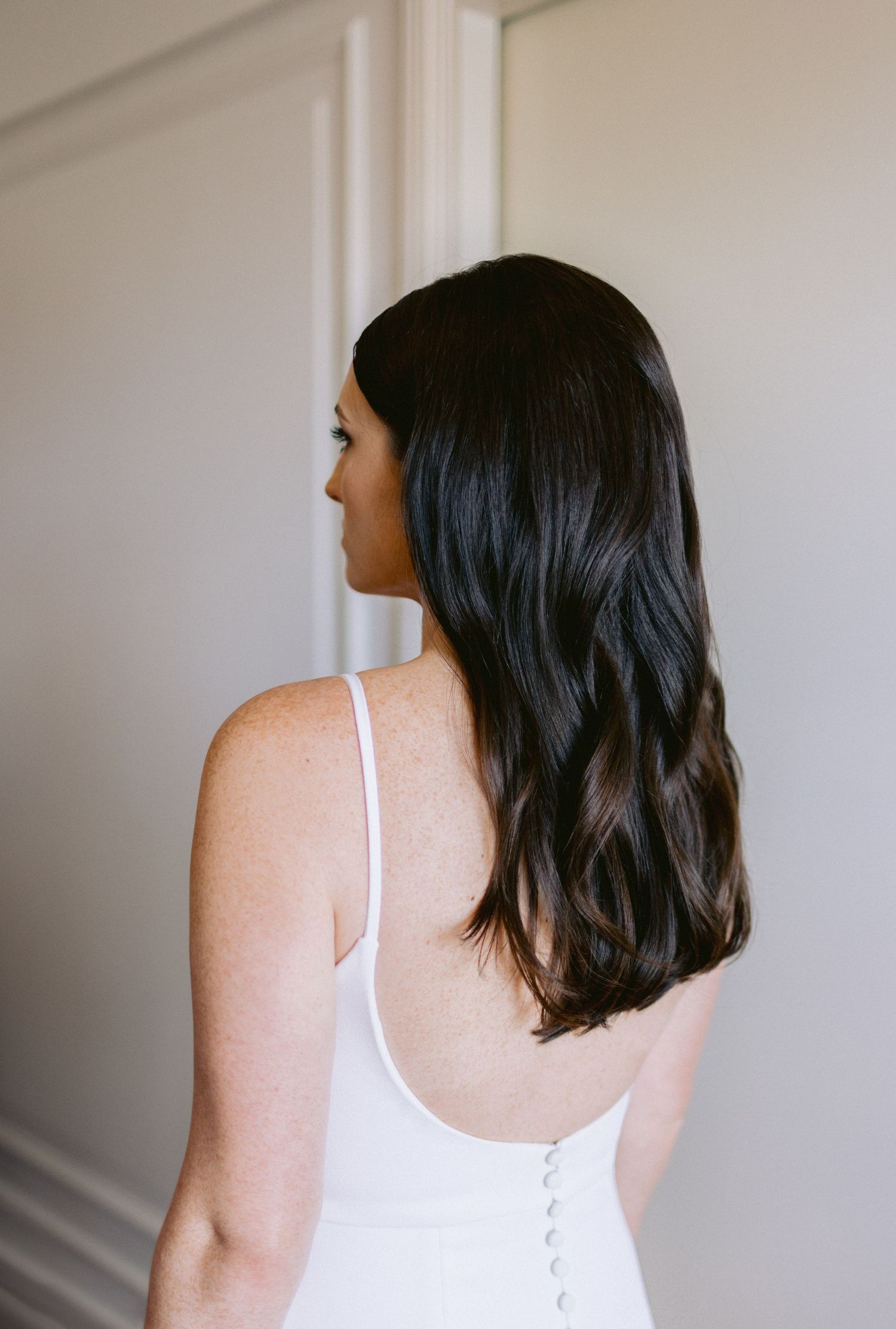 Minimalist bridal portrait showing back view in white dress against neutral wall.