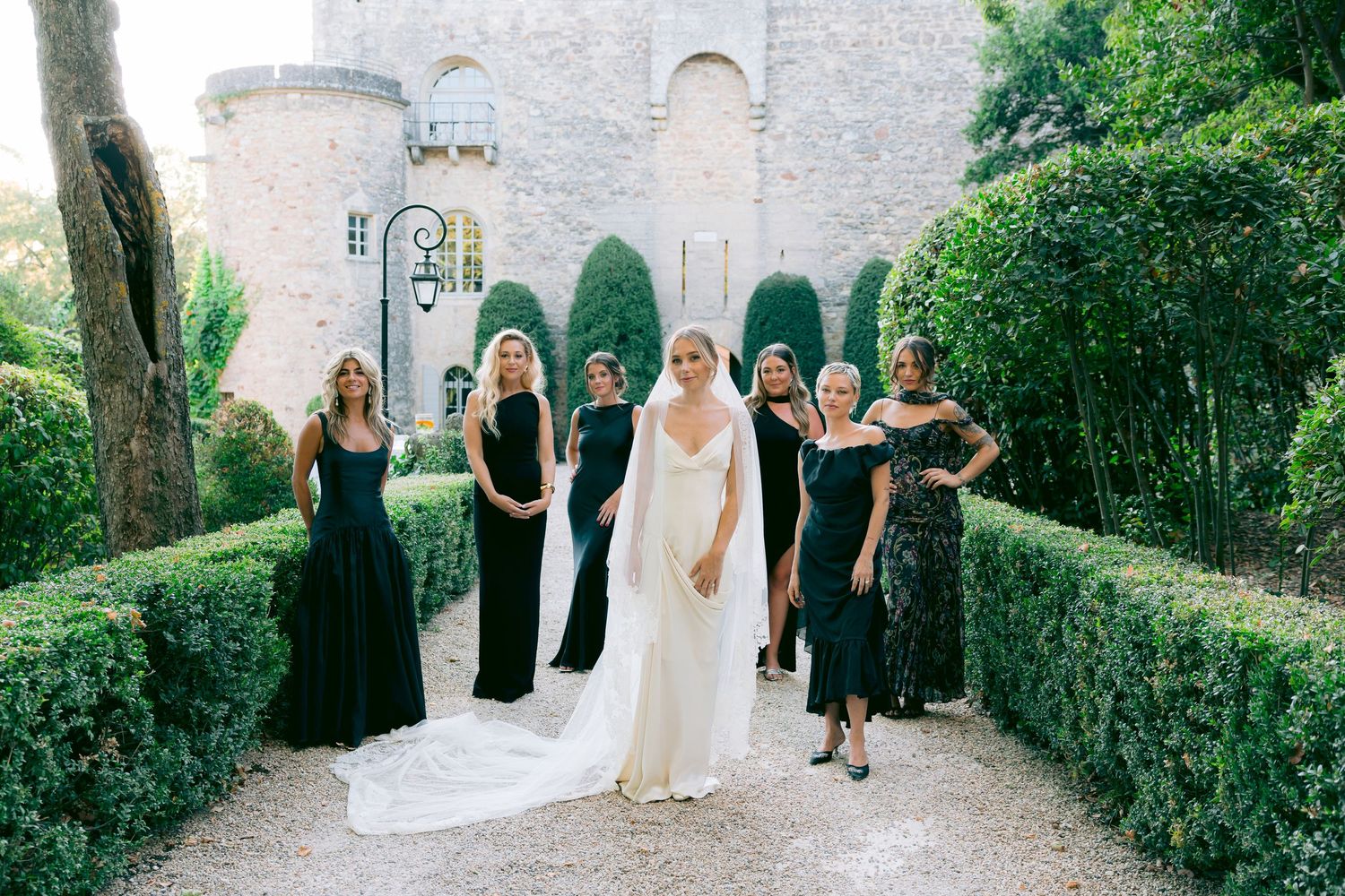 Wedding party walks along a stone path lined with manicured hedges and cypress trees.