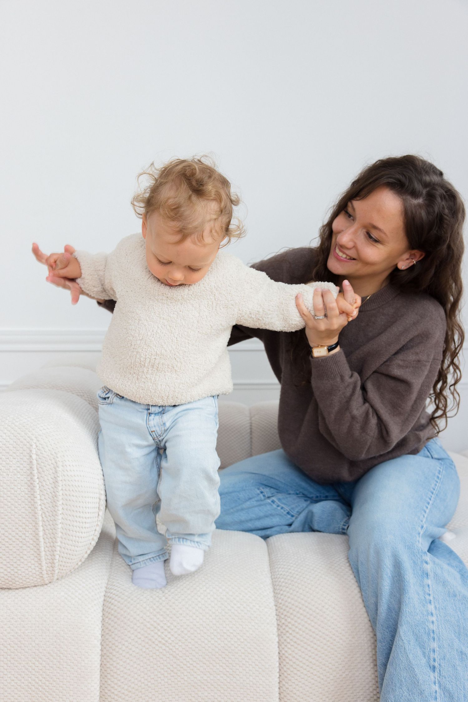 A smiling mother holds her toddler's hand as the baby stands on a white sofa, both wearing casual sweaters and jeans.