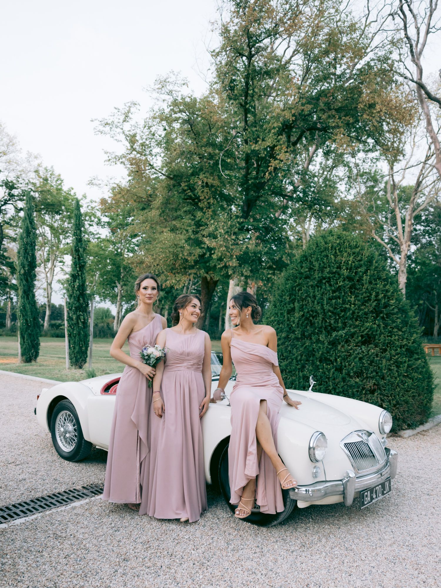 Three people in pink dresses pose with a classic white vintage MG sports car.