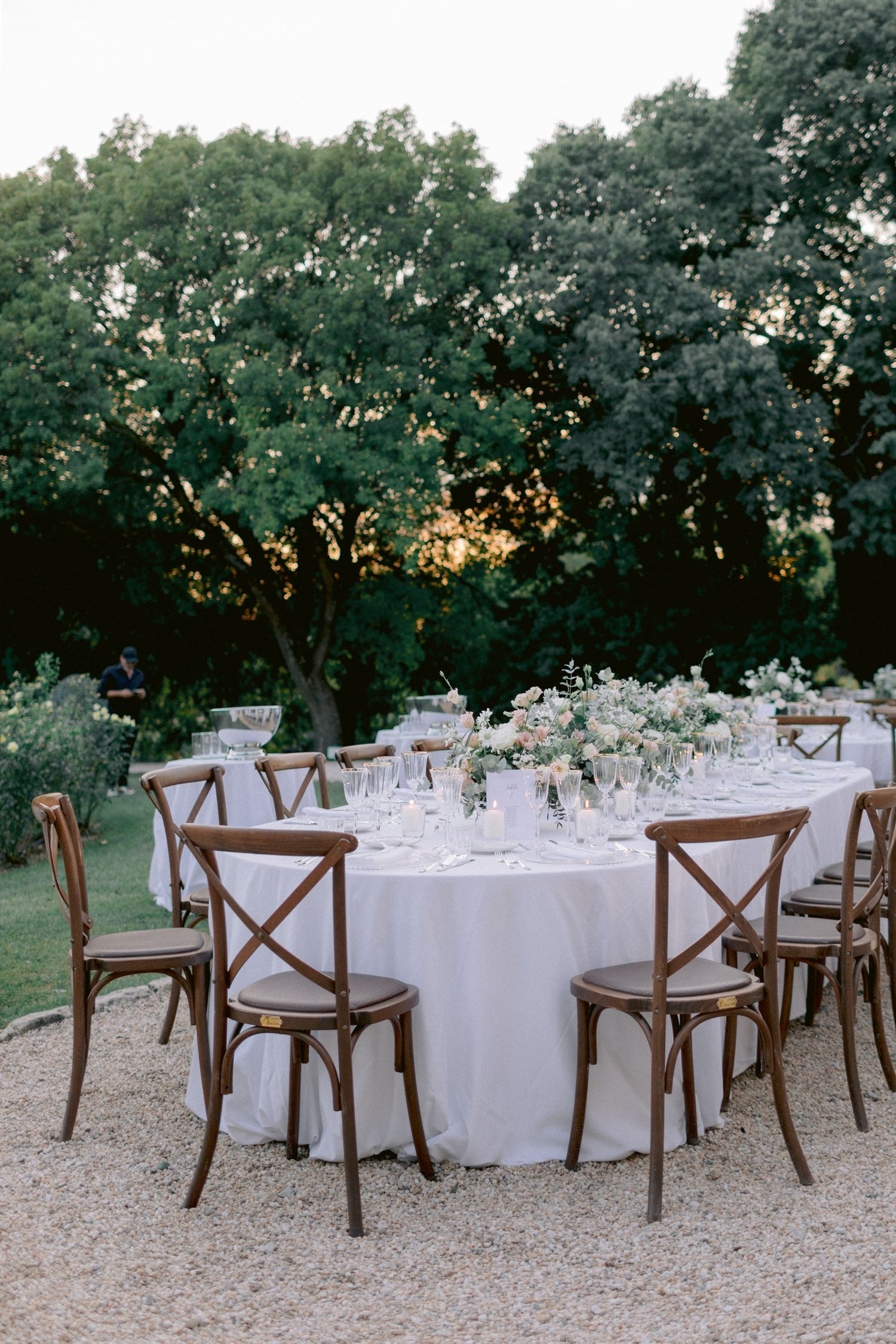Outdoor wedding reception setup with white tablecloths and wooden chairs arranged under trees at dusk.