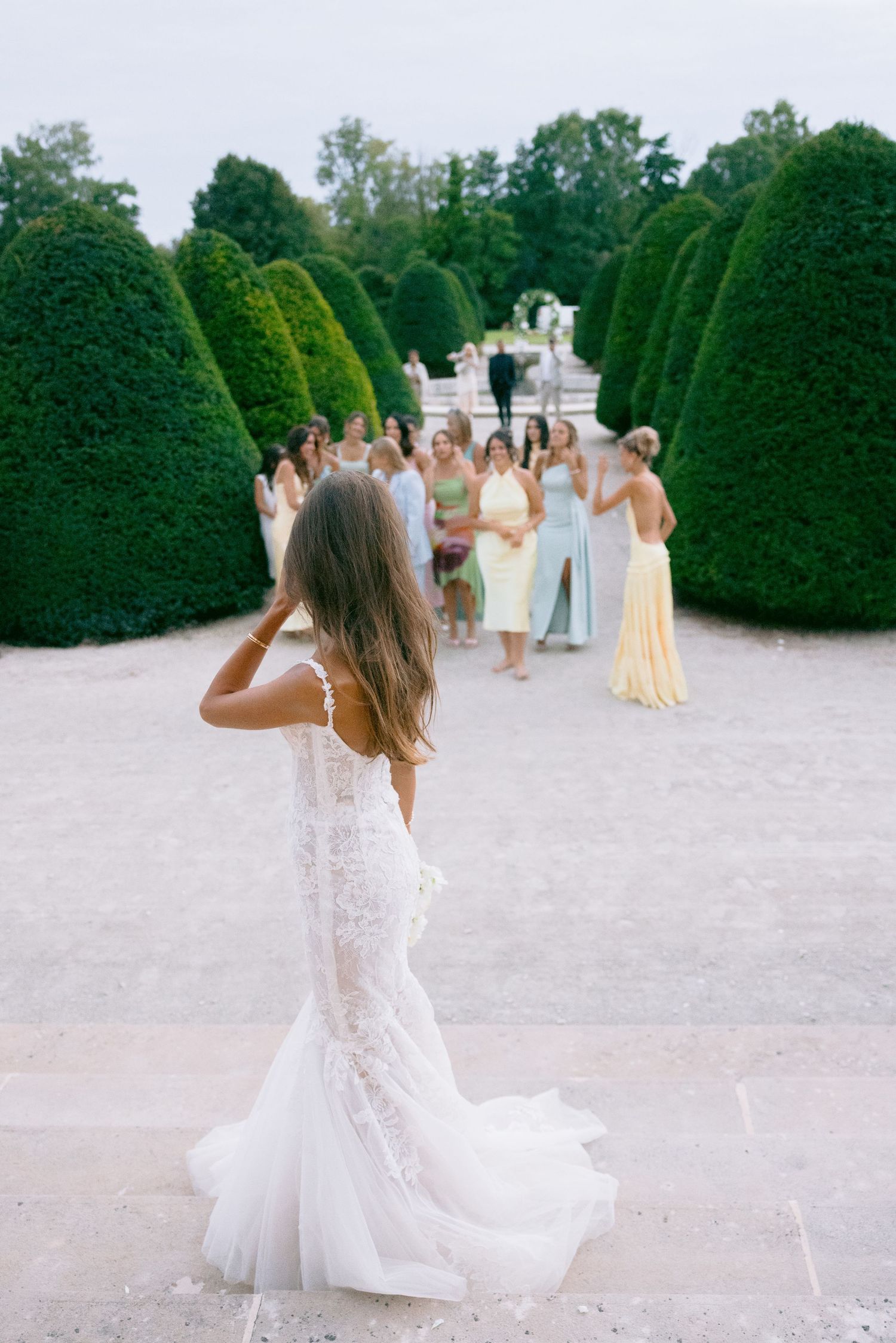 Bride in lace gown walks through formal garden with manicured hedges.