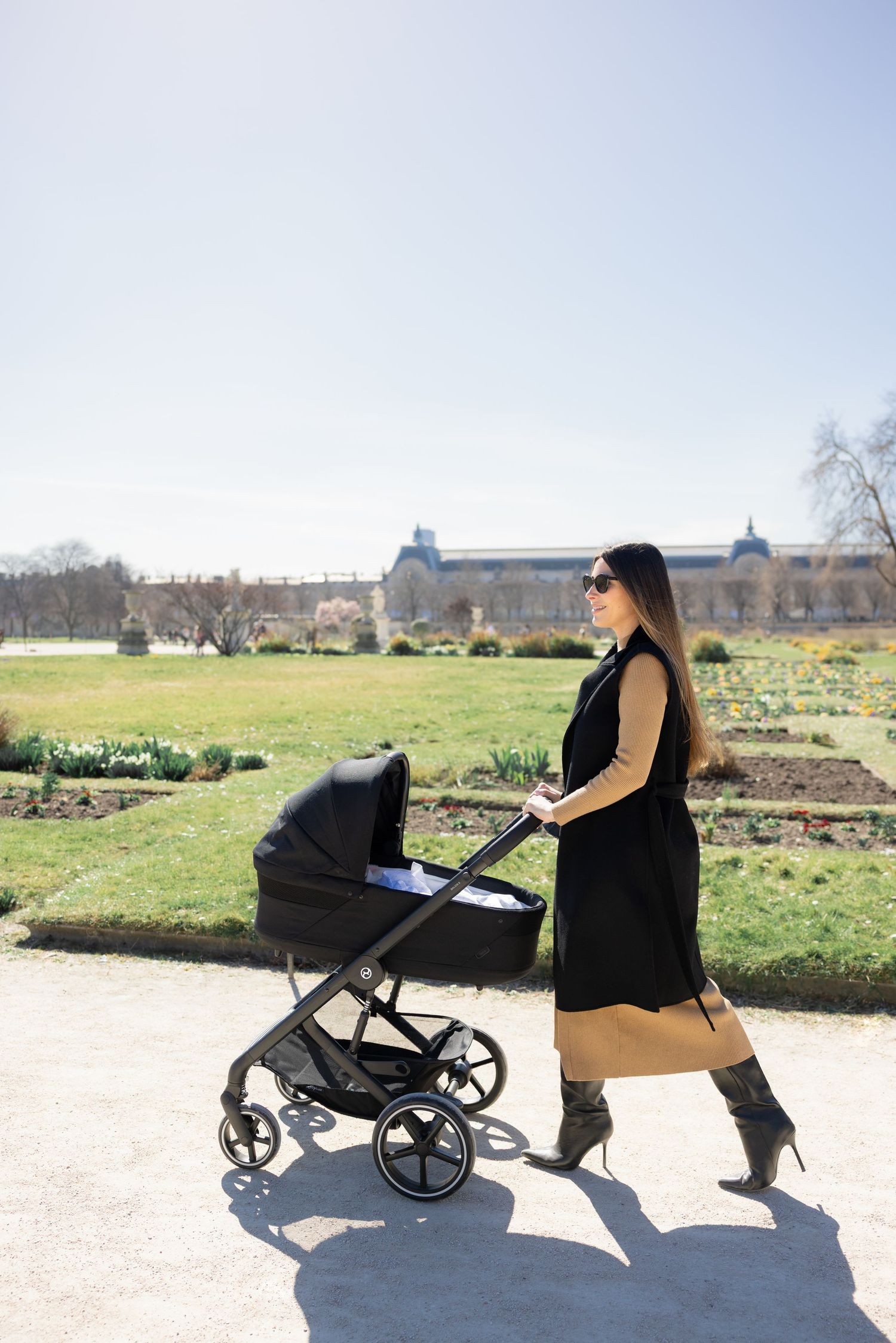 Woman pushing a modern black stroller along a sunny outdoor path in a garden setting.