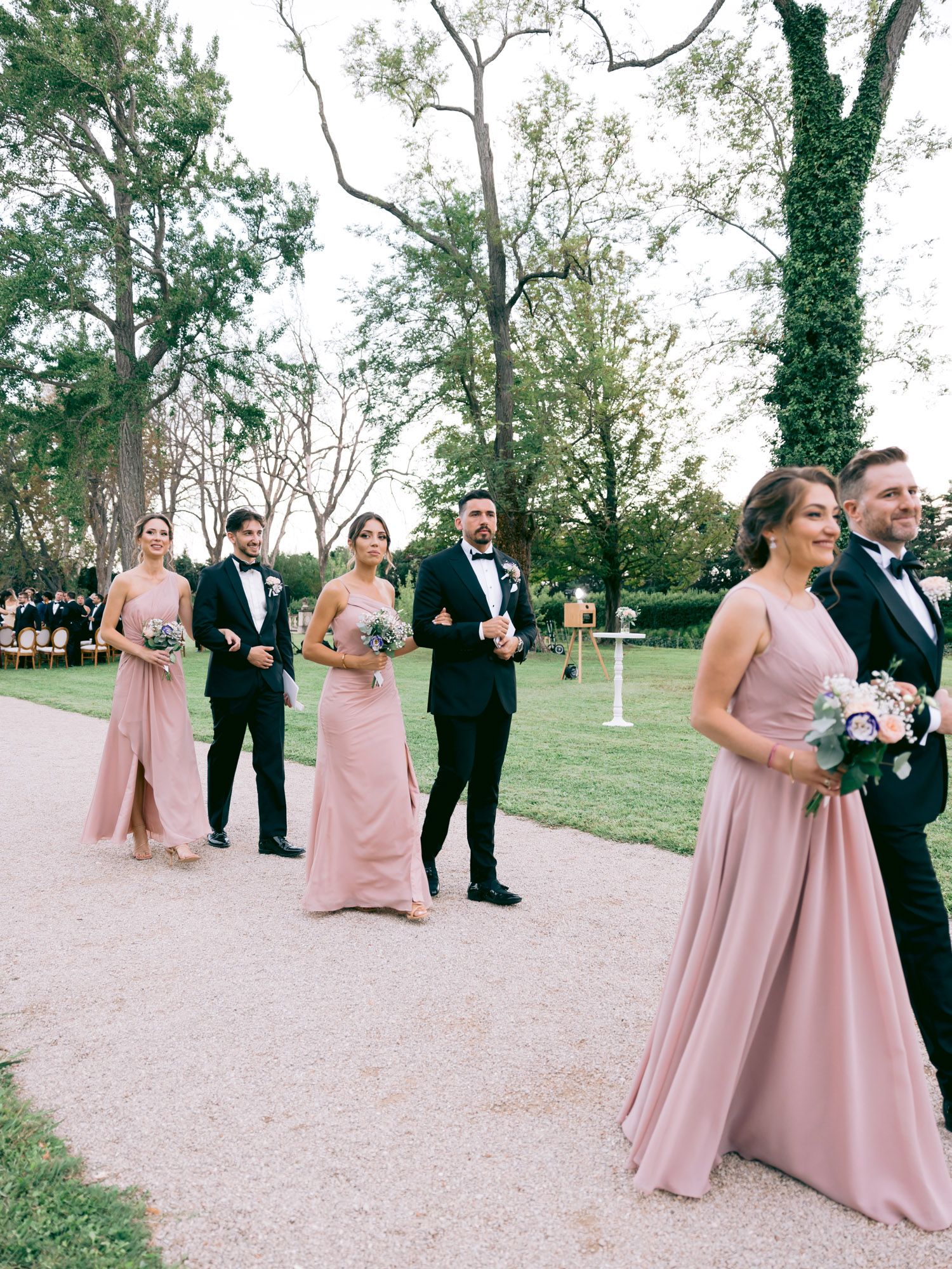 Wedding party members in pink gowns and black tuxedos walk along a garden path.