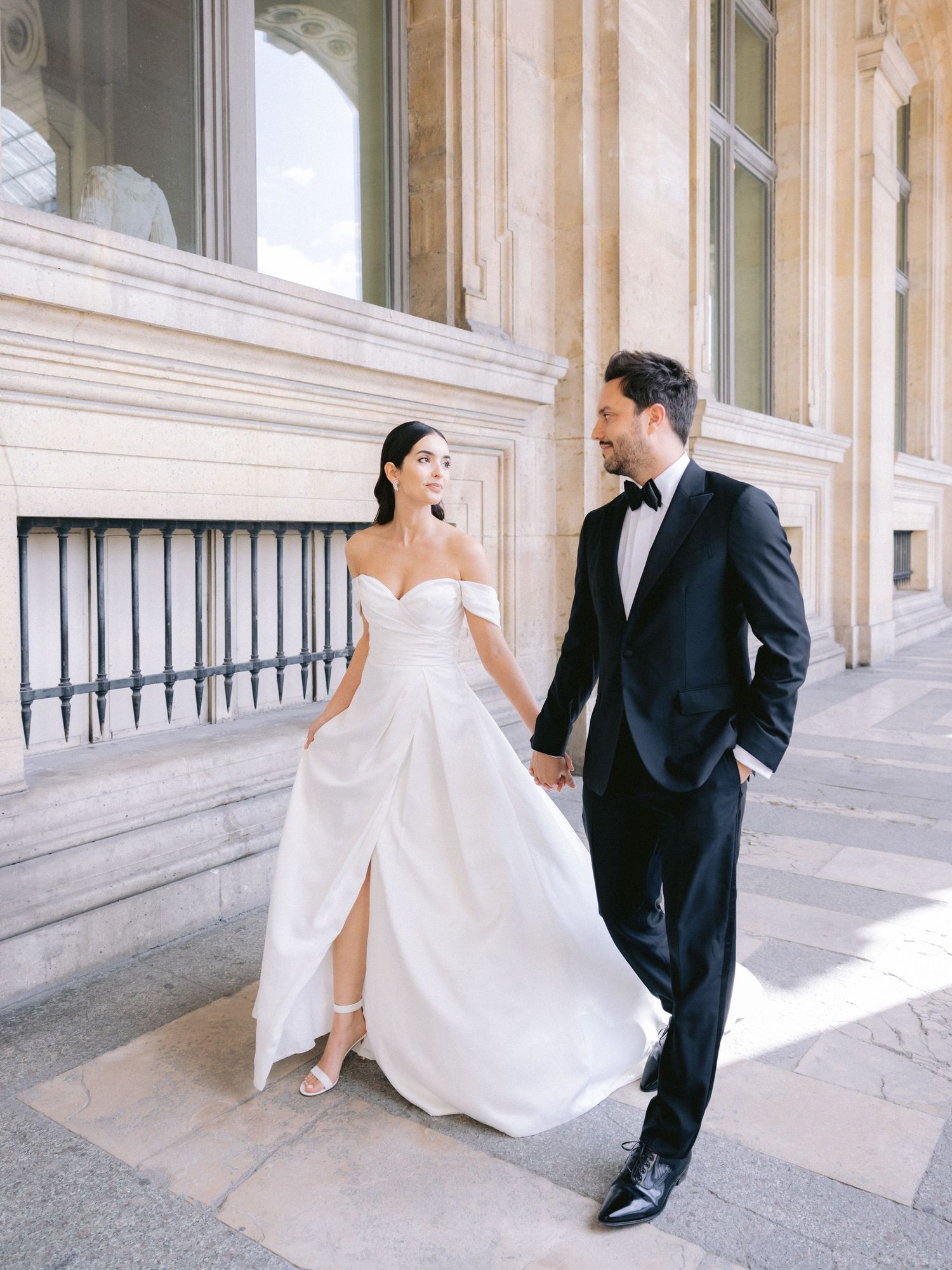 Newlyweds stroll through an elegant columned gallery with natural lighting.