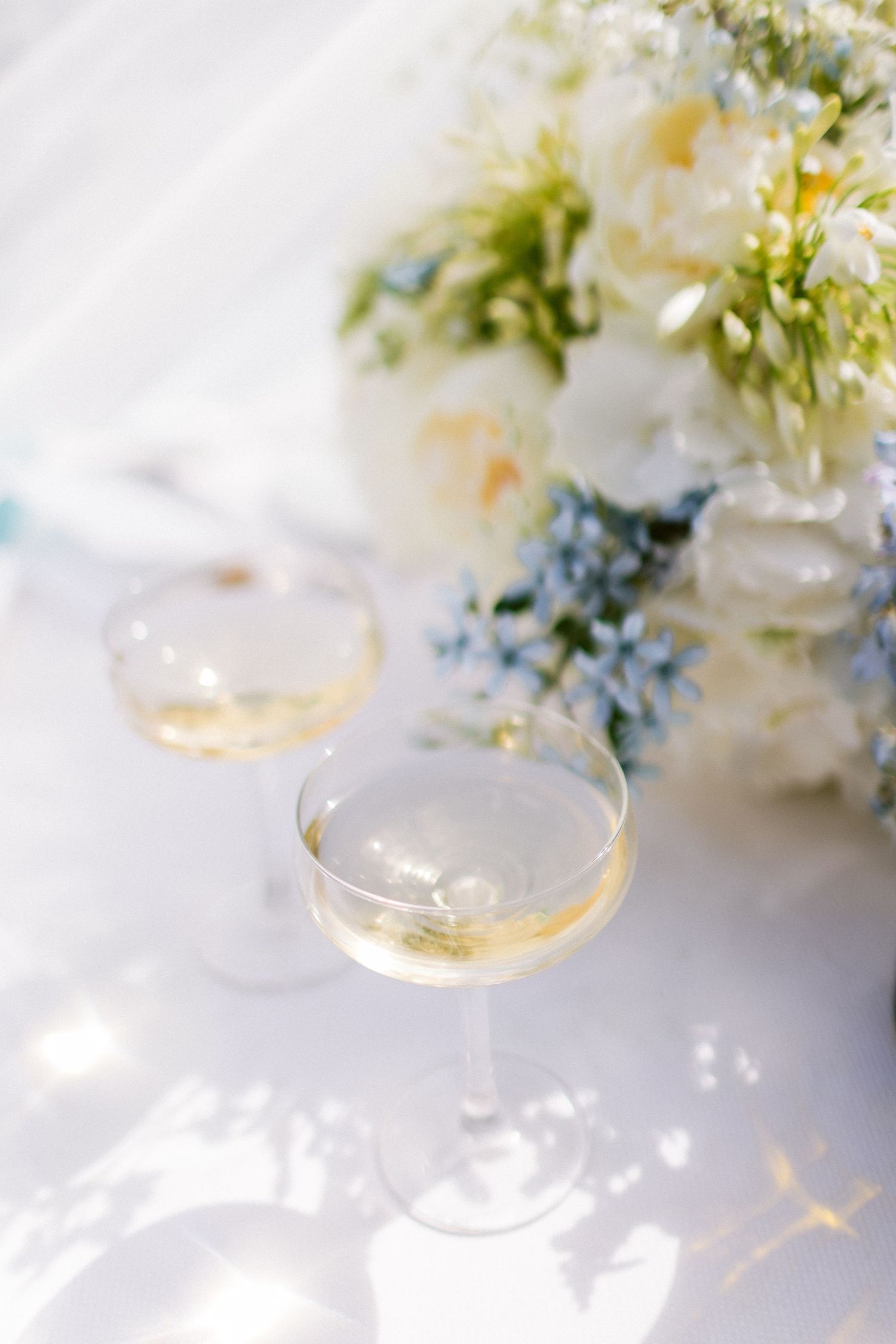 Delicate champagne glasses with white floral arrangements in soft natural light.