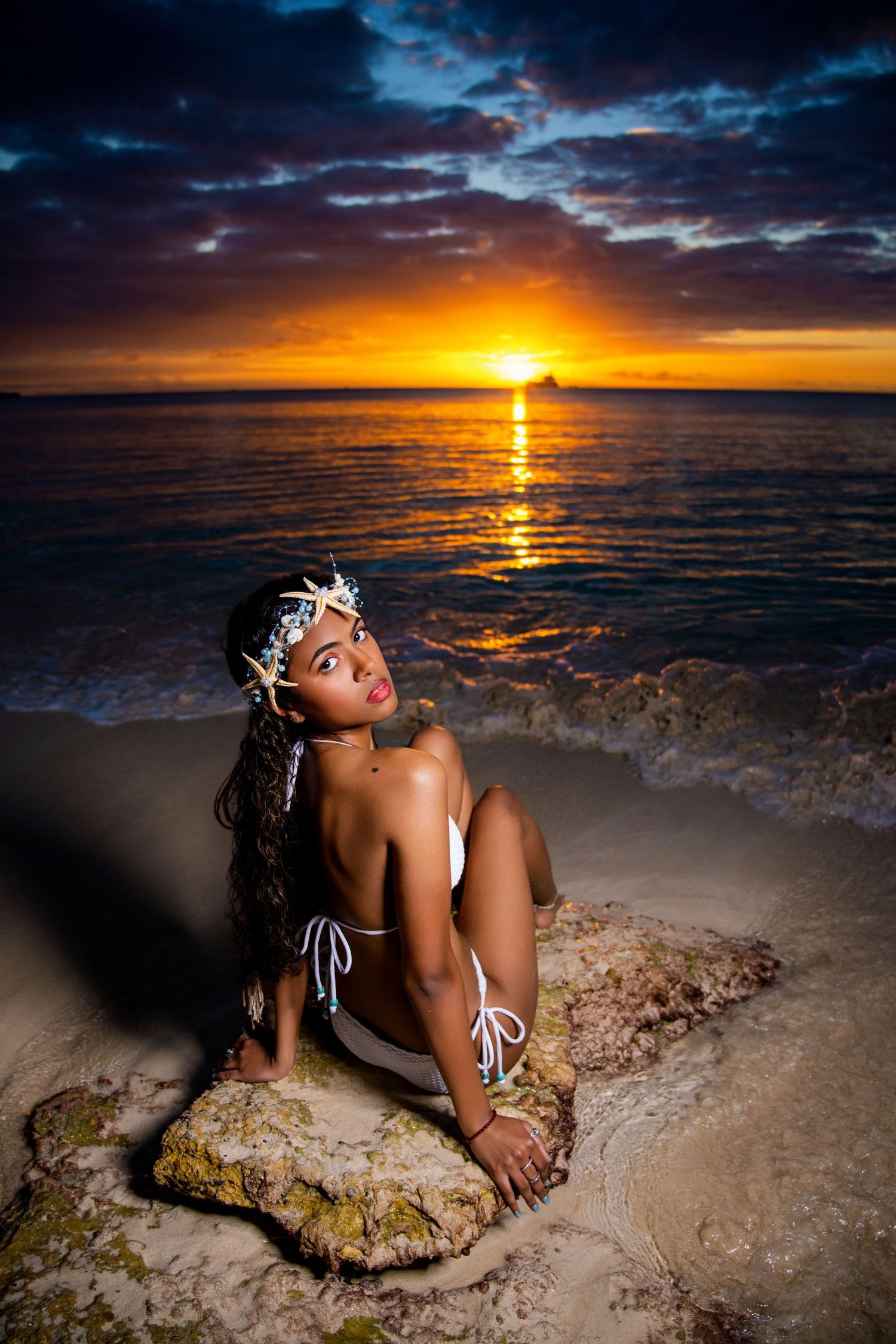 Dramatic beach sunset scene with figure posed on coastal rocks as golden sun dips below dark cloudy horizon in Antigua by Mark Blan.