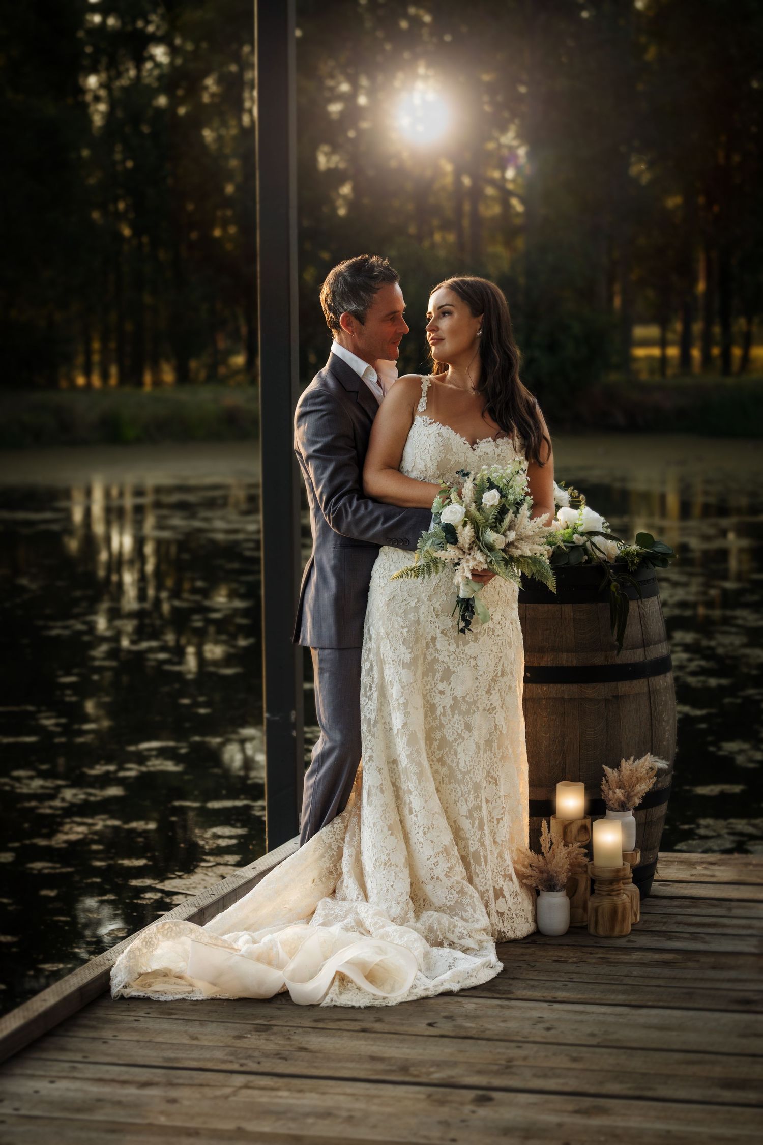 Romantic sunset wedding portrait with lace dress and rustic decor on wooden dock by tranquil pond.