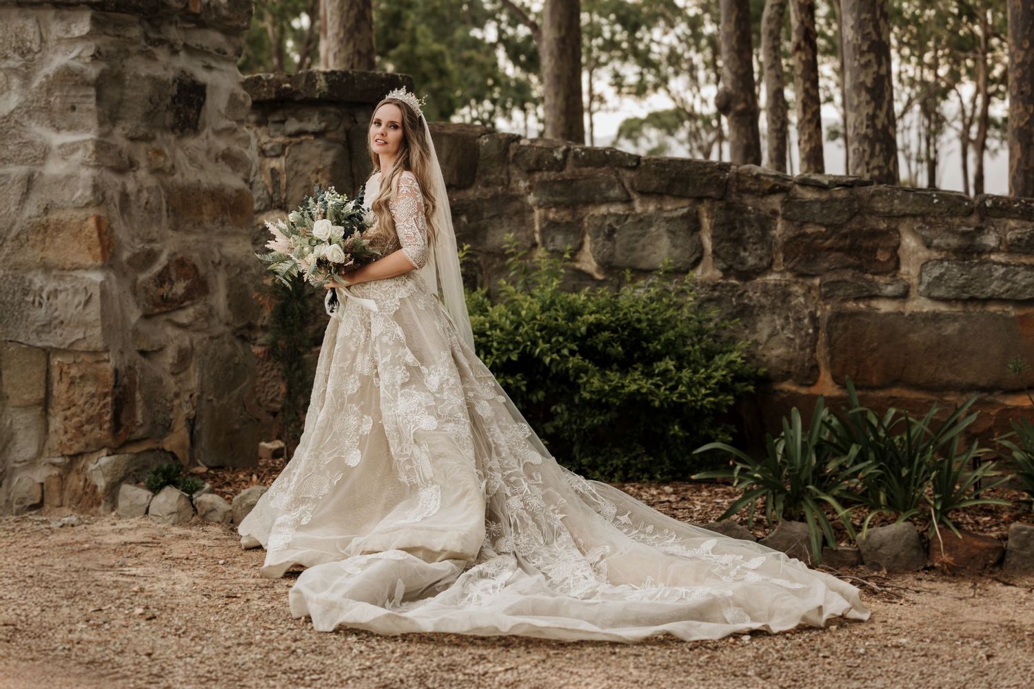 Elegant wedding dress with long lace train and bouquet photographed against rustic stone wall in forest setting.