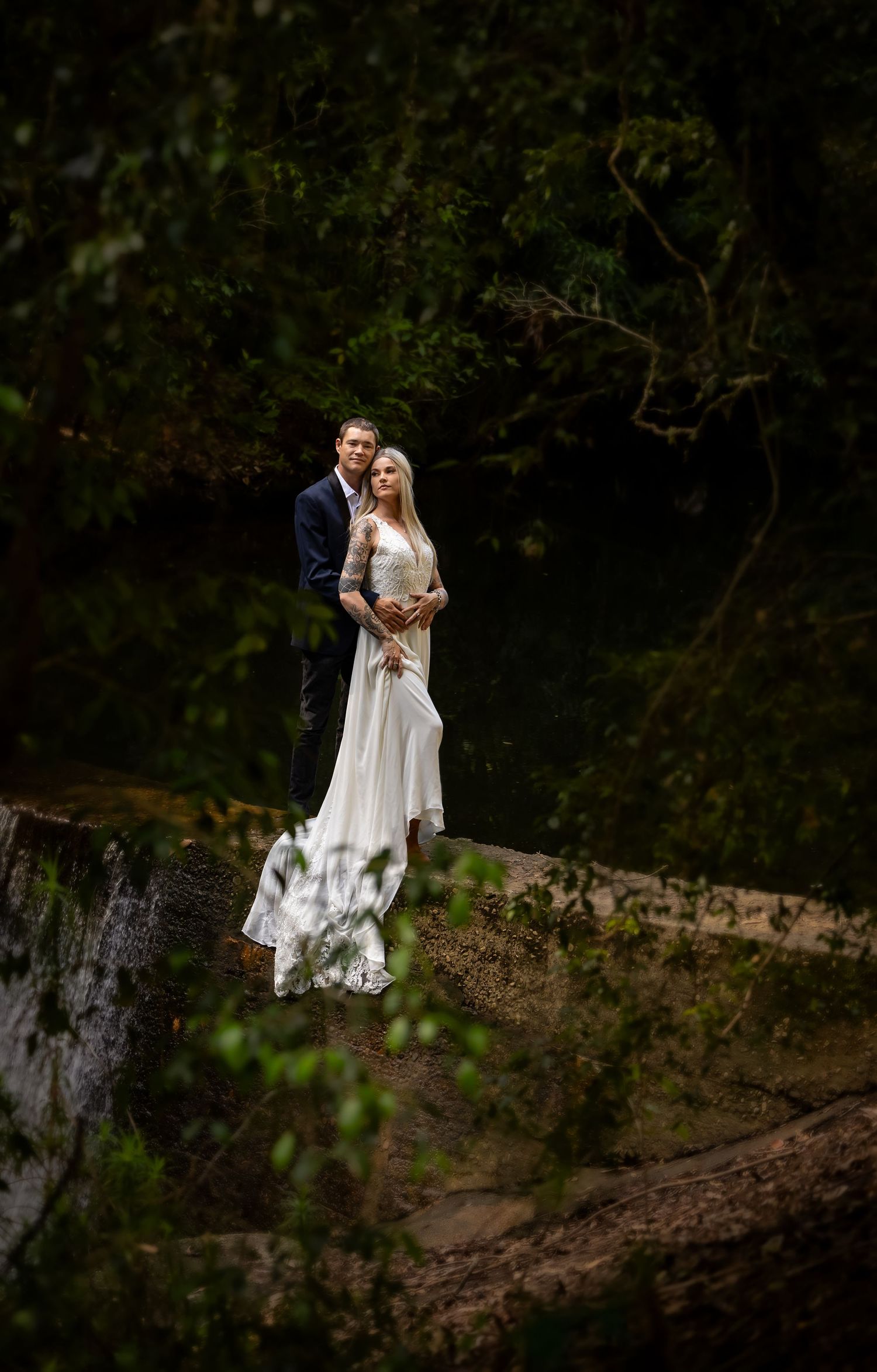 A wedding couple stands together on a hill against a dark forest backdrop in a moody, artistic composition.