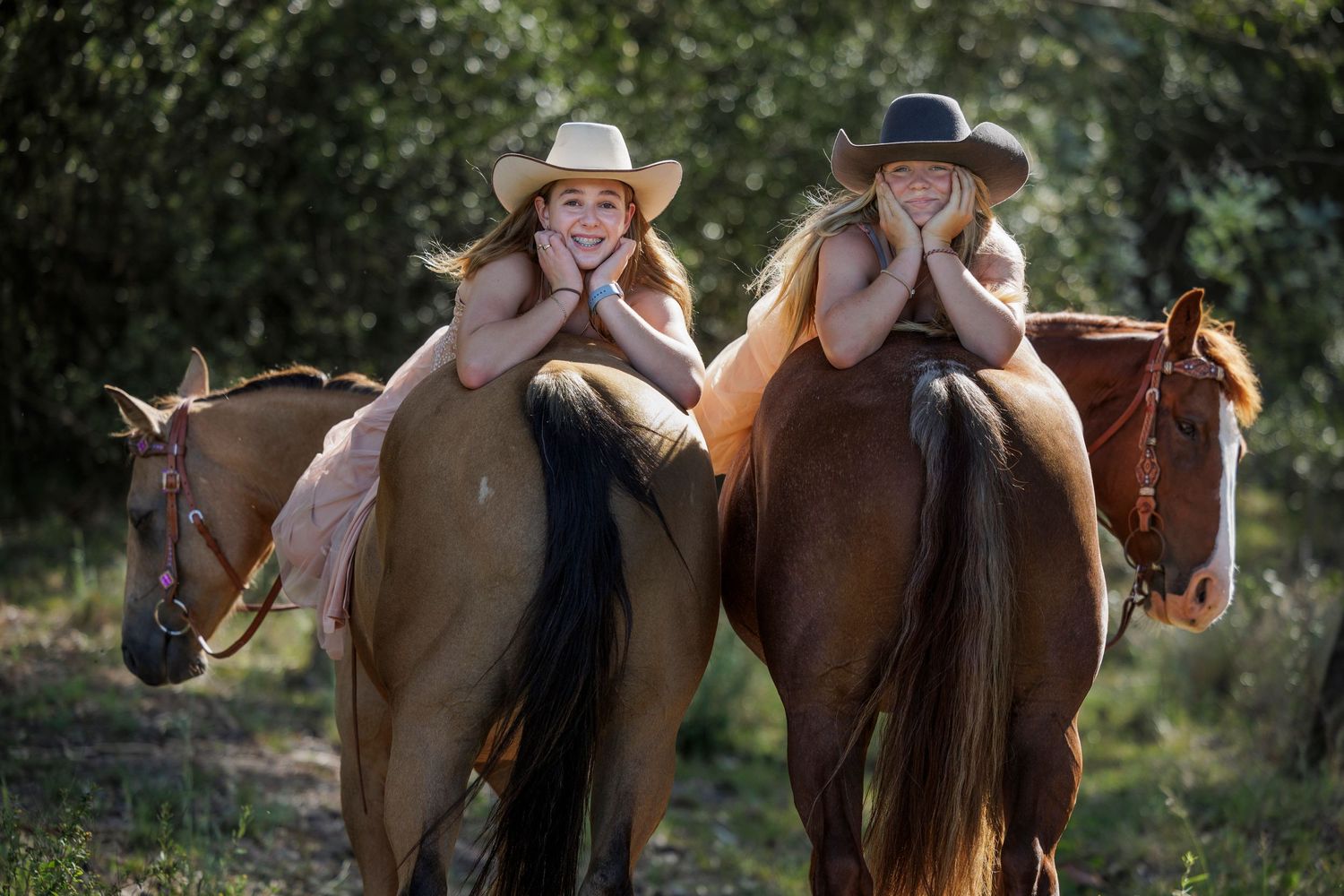Two equestrians in cowboy hats posing playfully on horseback in a sunlit forest.