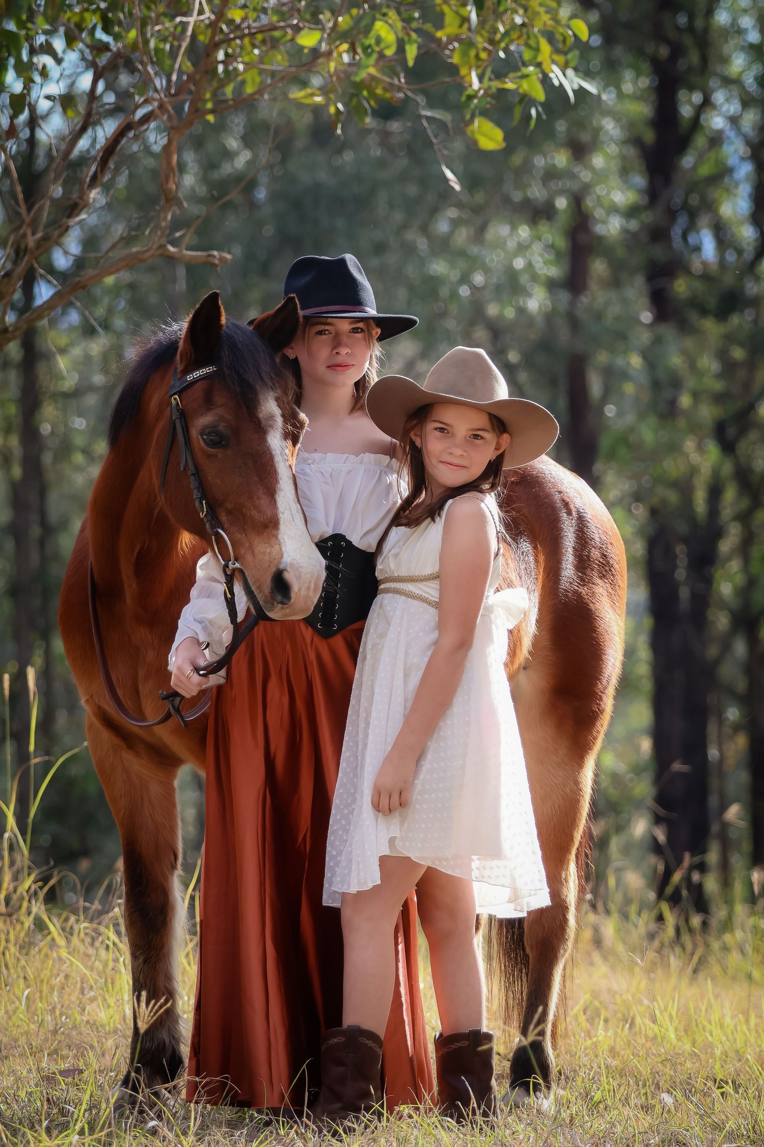A beautiful bay horse stands with two sisters wearing cowgirls hats in a rustic outdoor setting with trees and tall grass in the background.