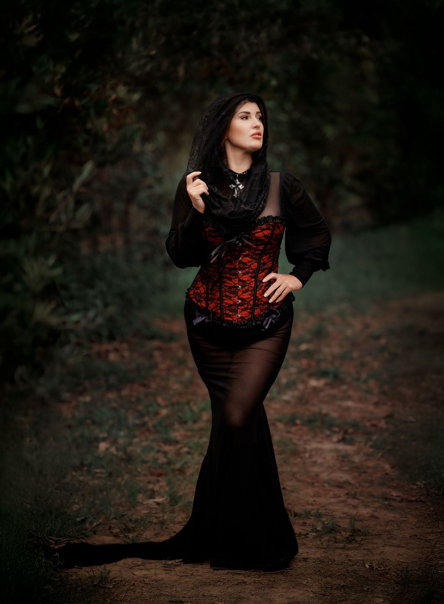 Gothic fashion model in red corset and black dress poses dramatically on forest path in moody portrait.
