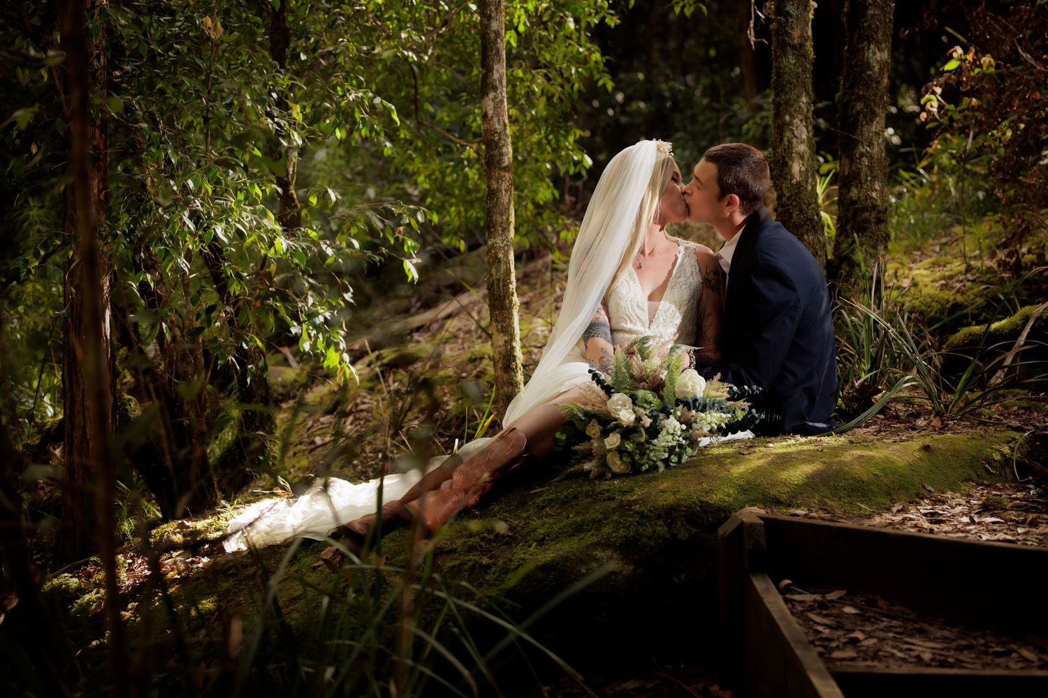 A romantic wedding couple sits together on mossy rocks in a serene forest setting with soft natural lighting.