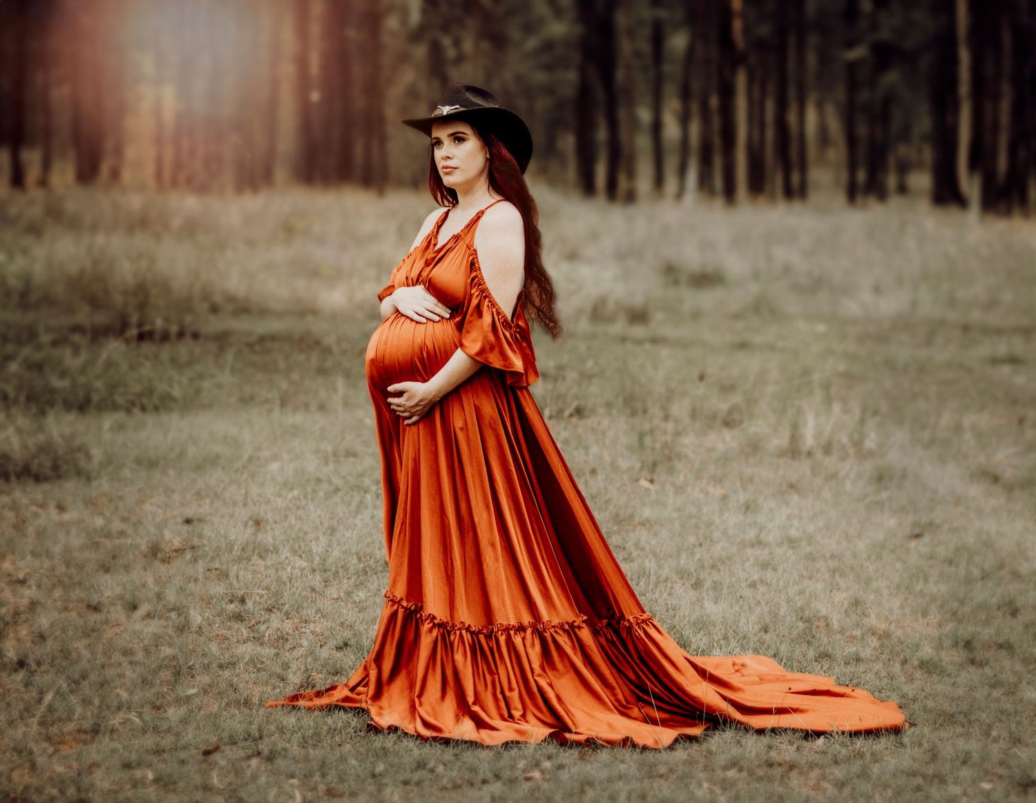 An expectant woman in a flowing burnt orange gown stands in a sunlit forest clearing.