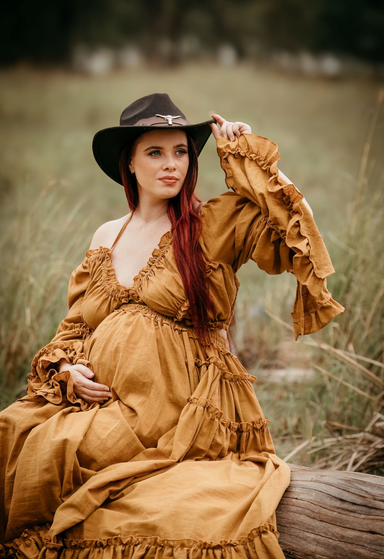 Rustic maternity portrait in a mustard prairie dress and wide-brim hat amid tall grass.