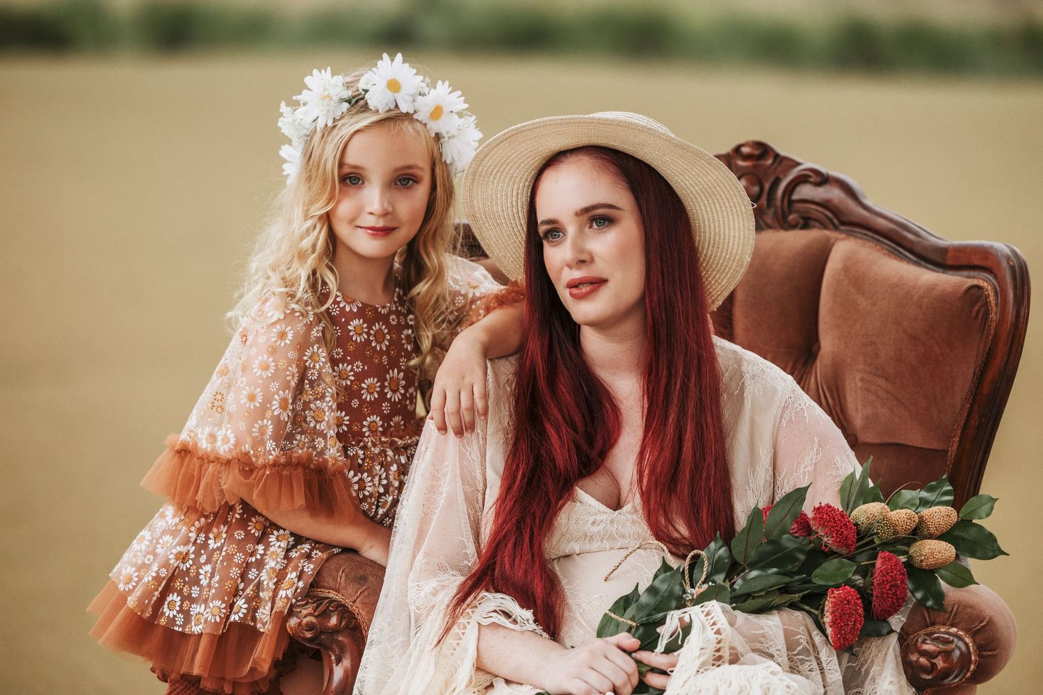 Two people pose in vintage-inspired outfits with floral crown and brown velvet chair in outdoor setting.