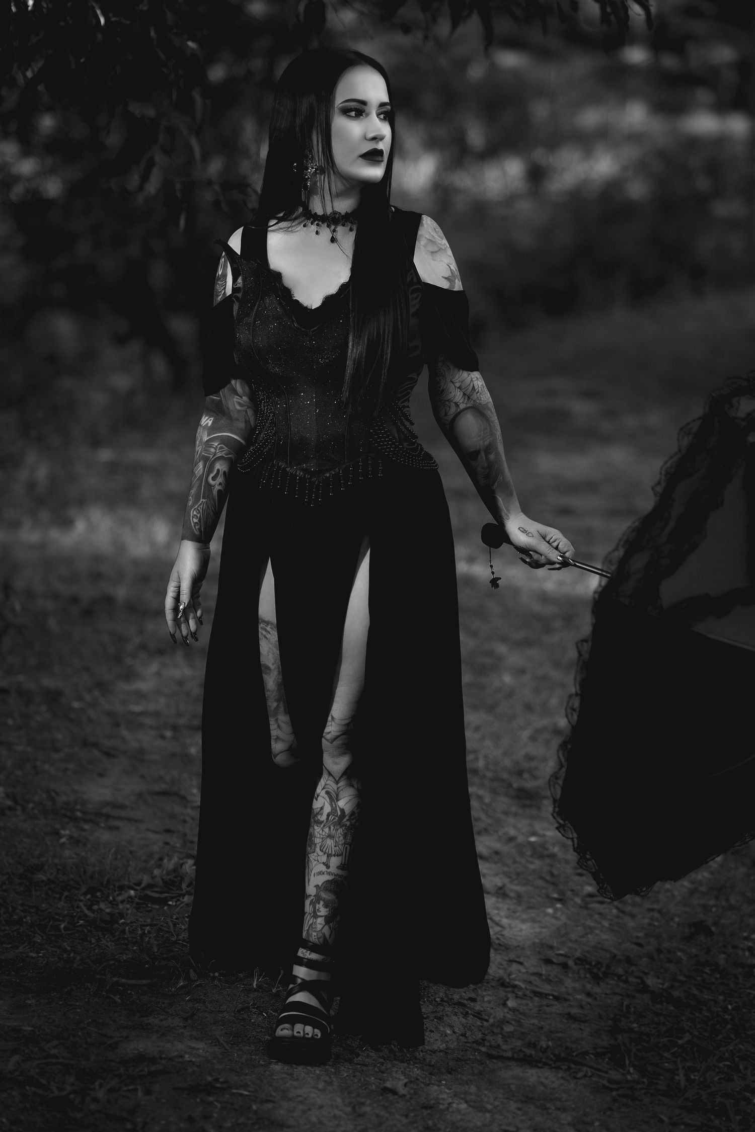 A gothic figure in a flowing black dress poses dramatically in a dark outdoor setting with moody lighting.