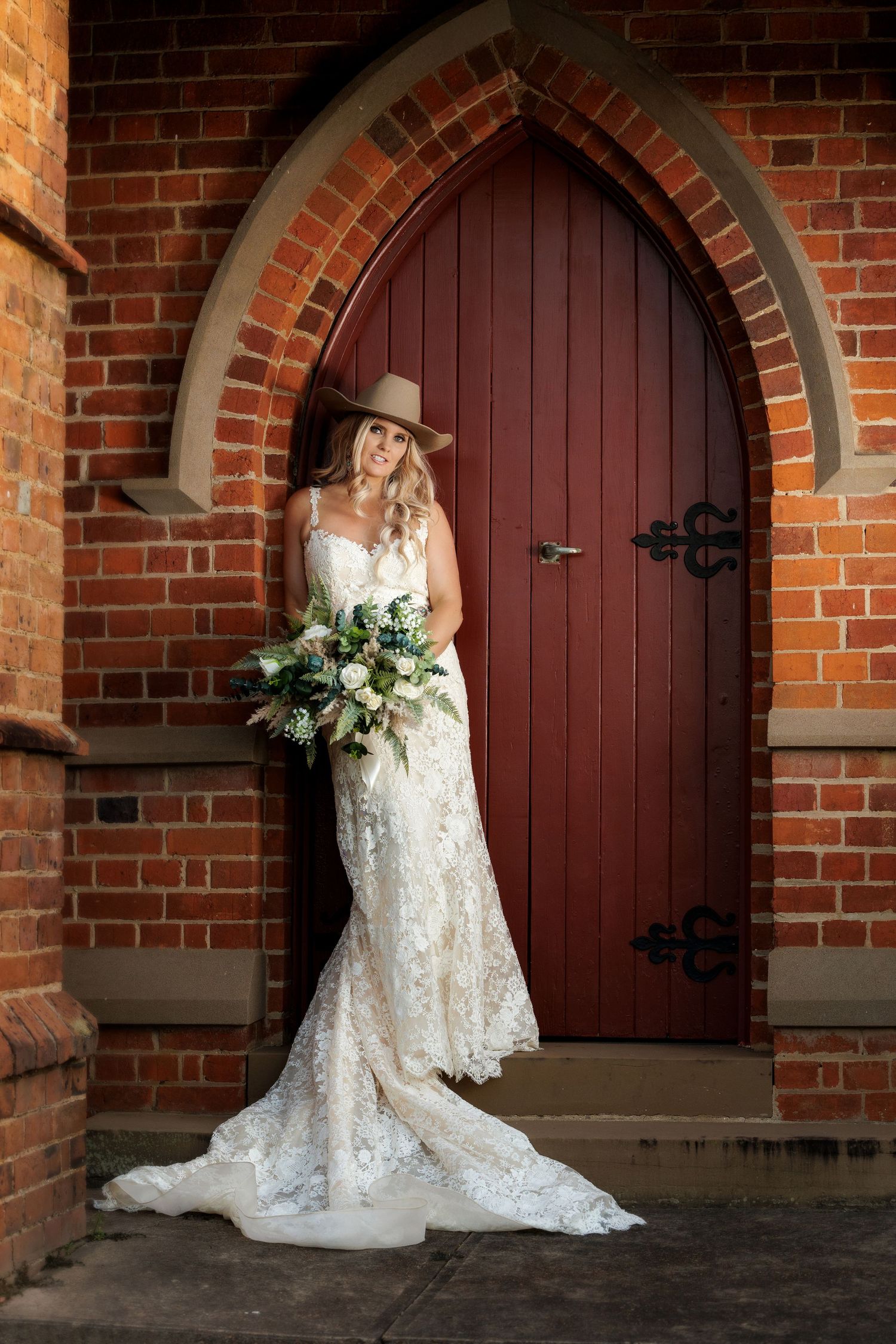 Elegant wedding portrait featuring white lace gown and bouquet against rustic brown wooden church door and brick archway.
