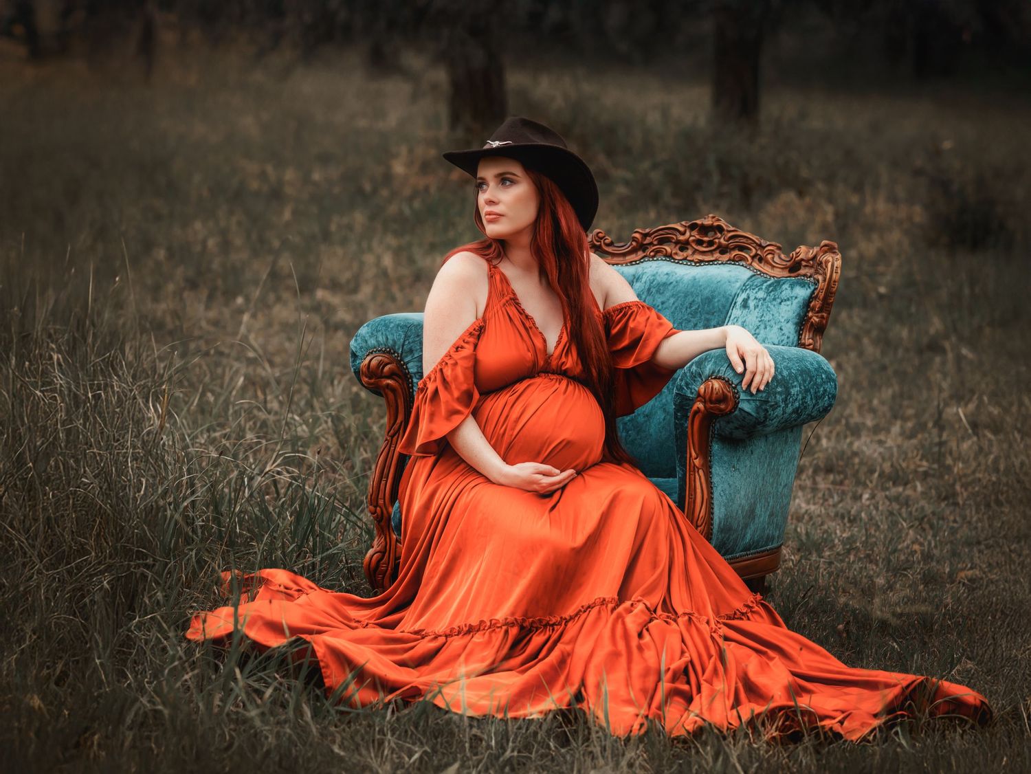 A person in a flowing orange dress sits on an antique turquoise armchair in a misty field.