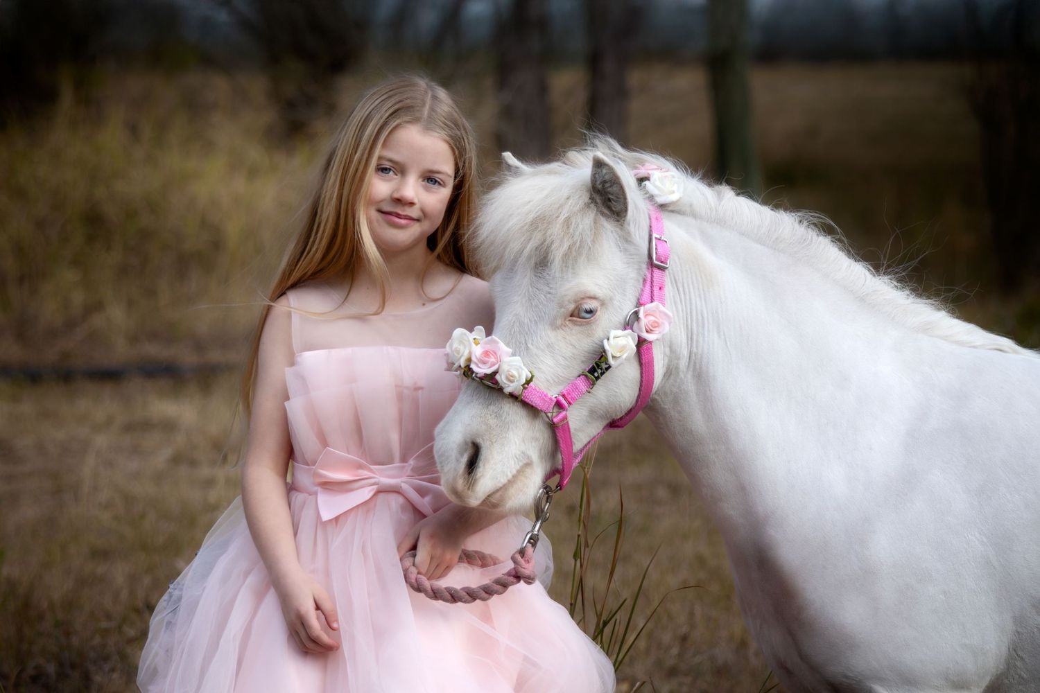 A white pony with pink accessories nuzzles close to someone in a pink dress.