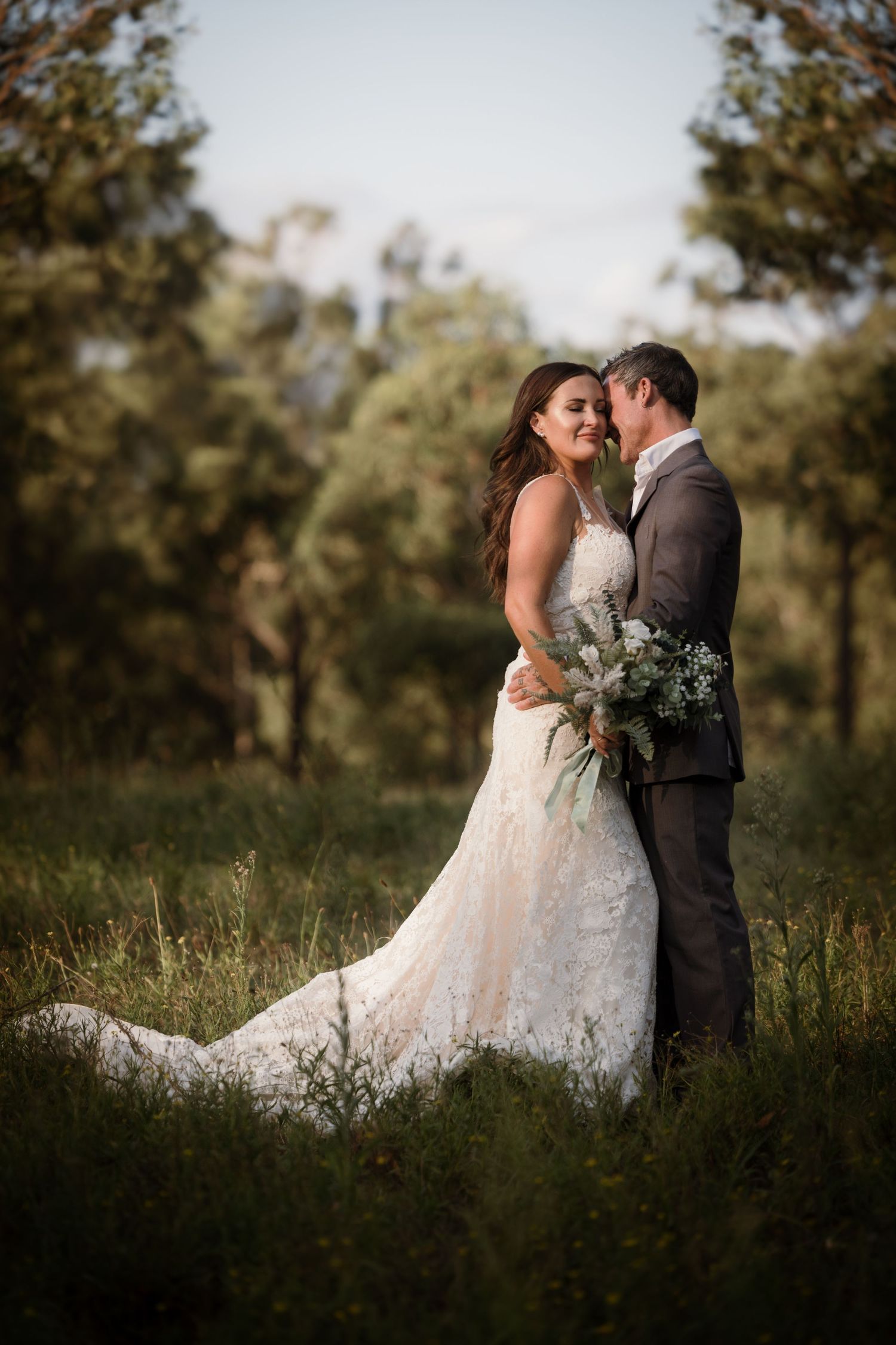 A romantic wedding couple embraces in a sunlit forest clearing while the bride holds a beautiful eucalyptus bouquet.