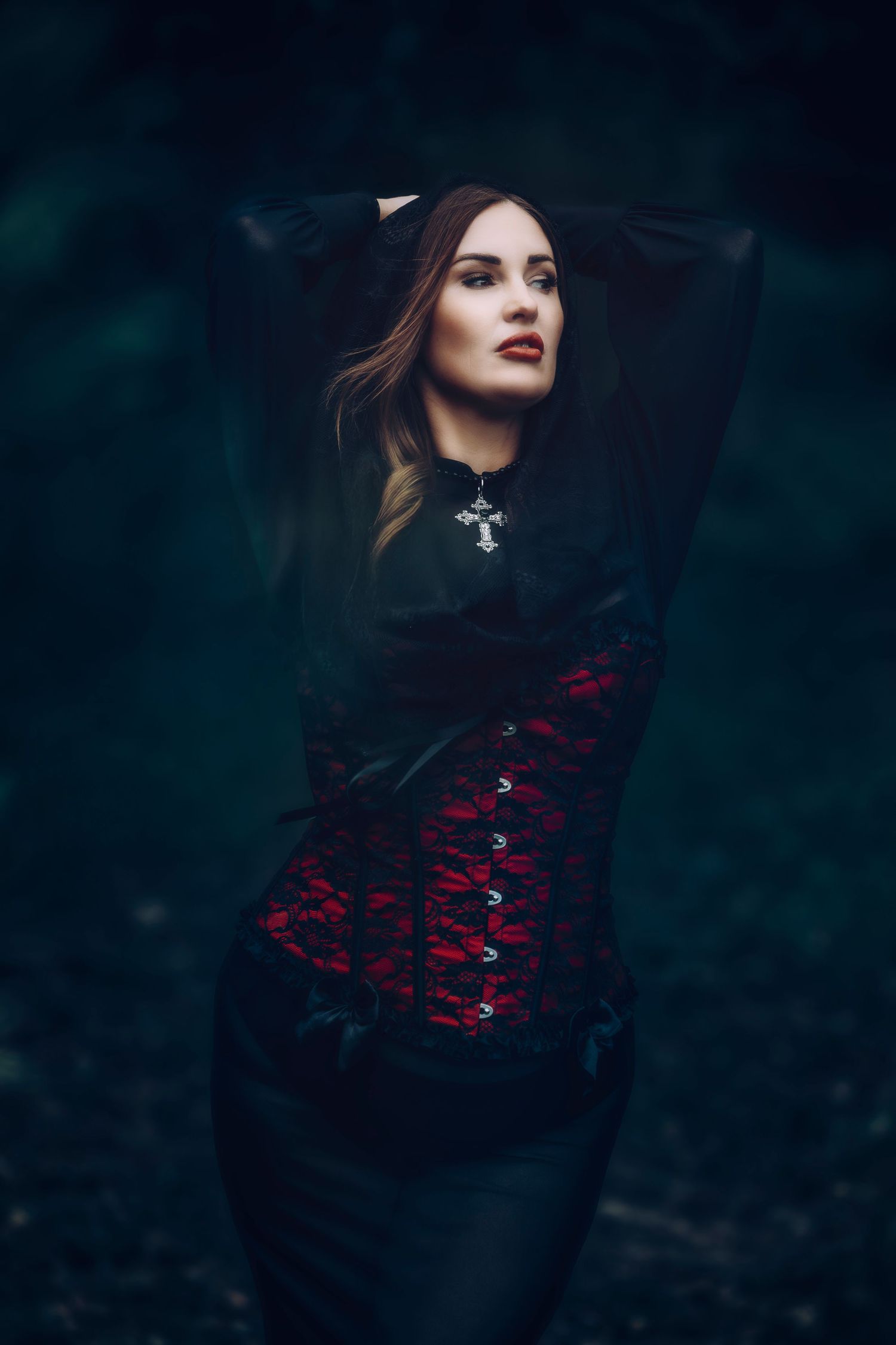 A figure in gothic fashion wearing a black dress and red corset poses dramatically against a dark moody background.