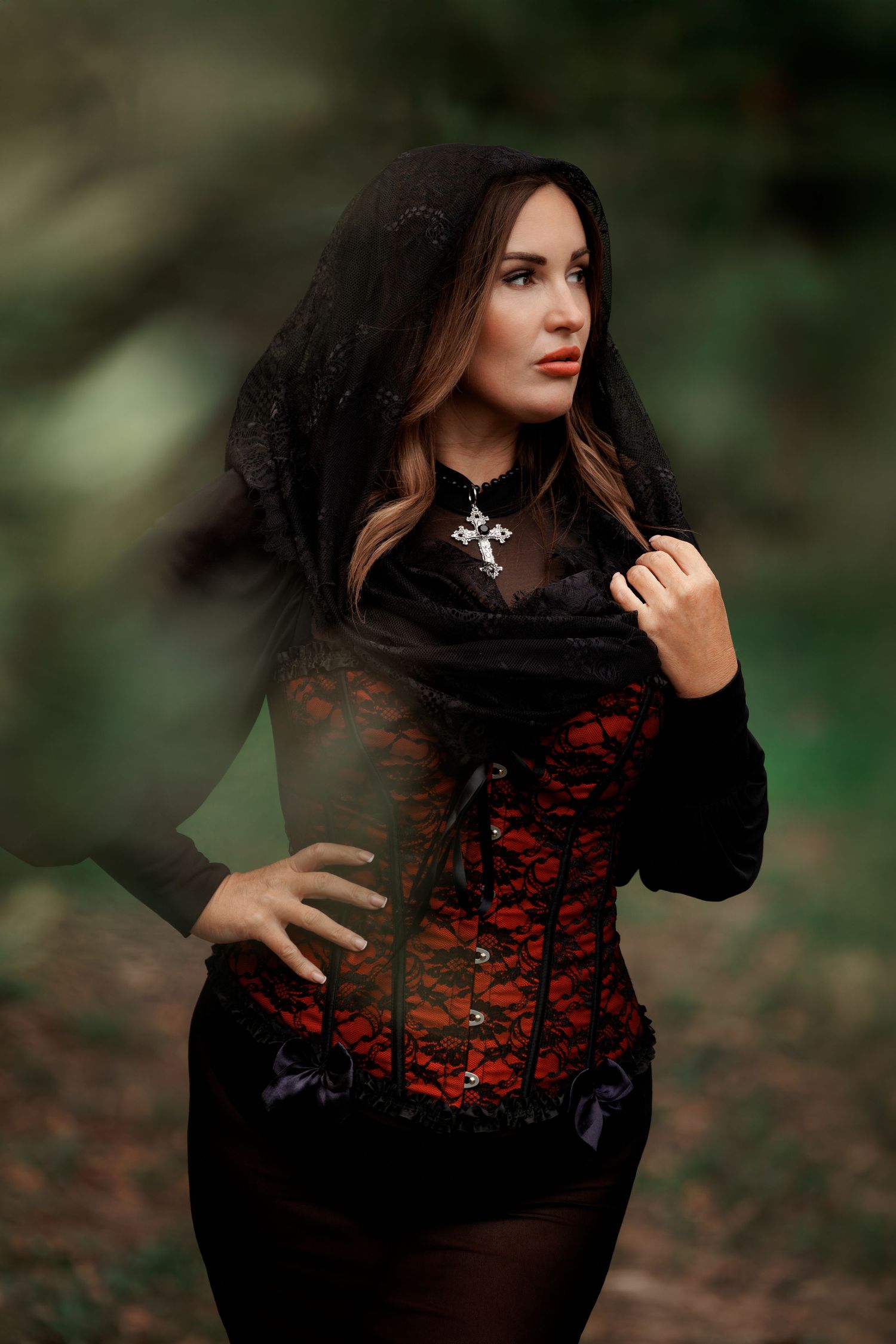 A gothic-styled portrait featuring a black hooded cape and red lace corset in a moody forest setting.