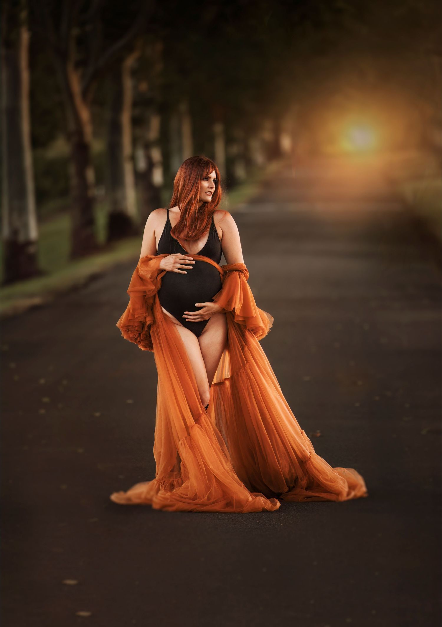 A pregnant woman in a flowing orange dress poses on a tree-lined path at sunset with dramatic lighting.