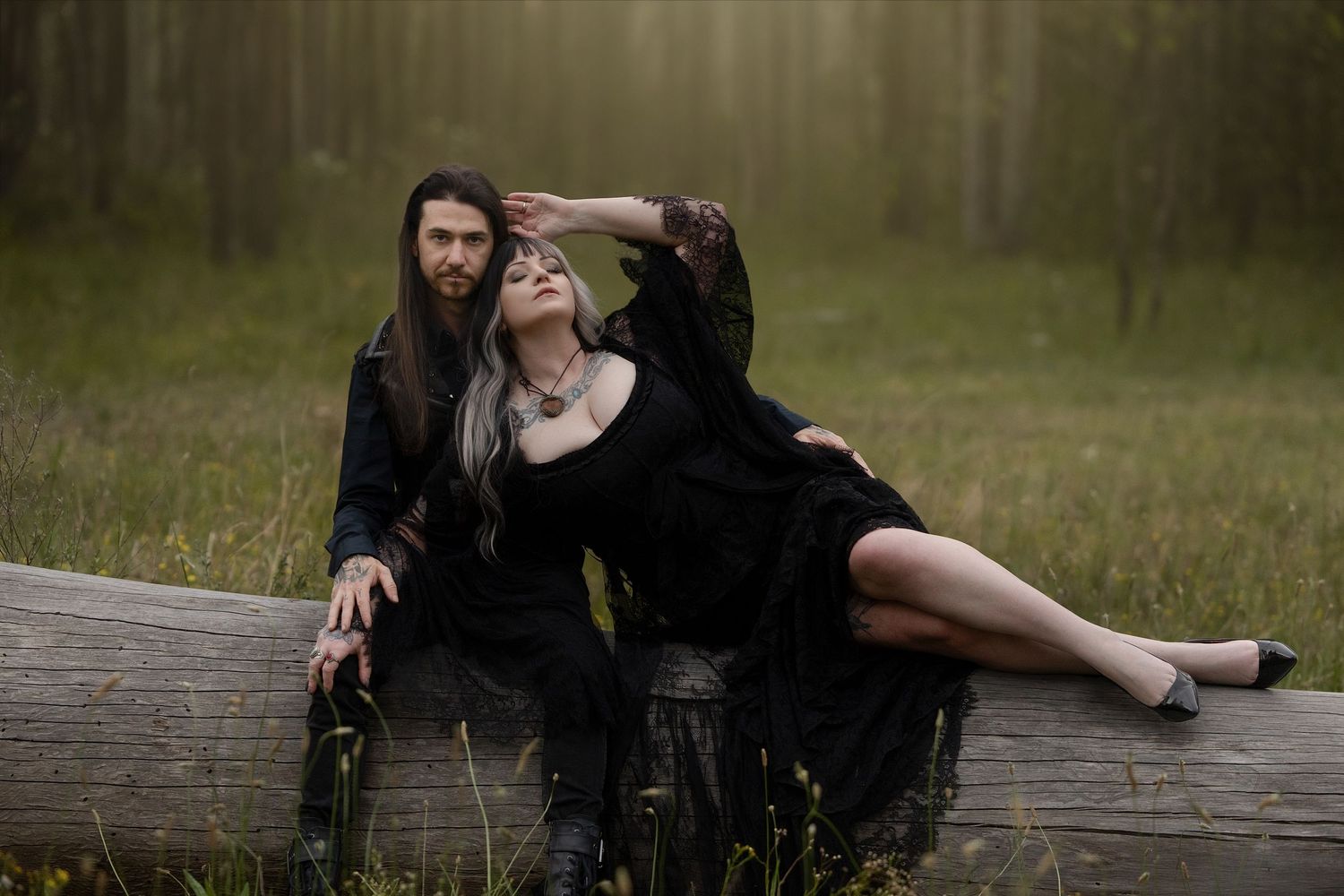 A person in dark clothing poses dramatically on a wooden fence in a grassy outdoor setting.