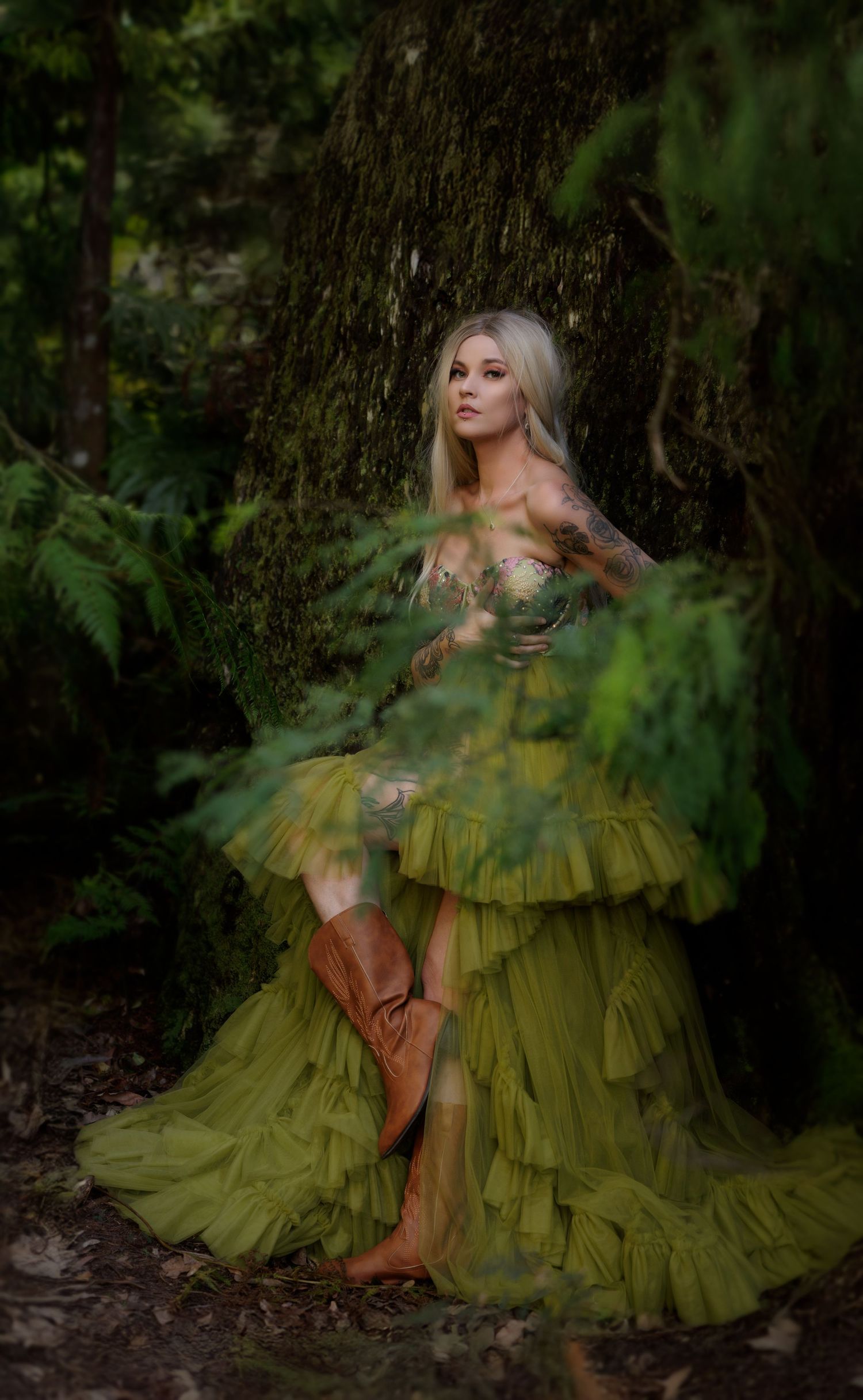 A figure in a flowing green ruffled gown poses ethereally against a mossy tree trunk in a mystical forest setting.