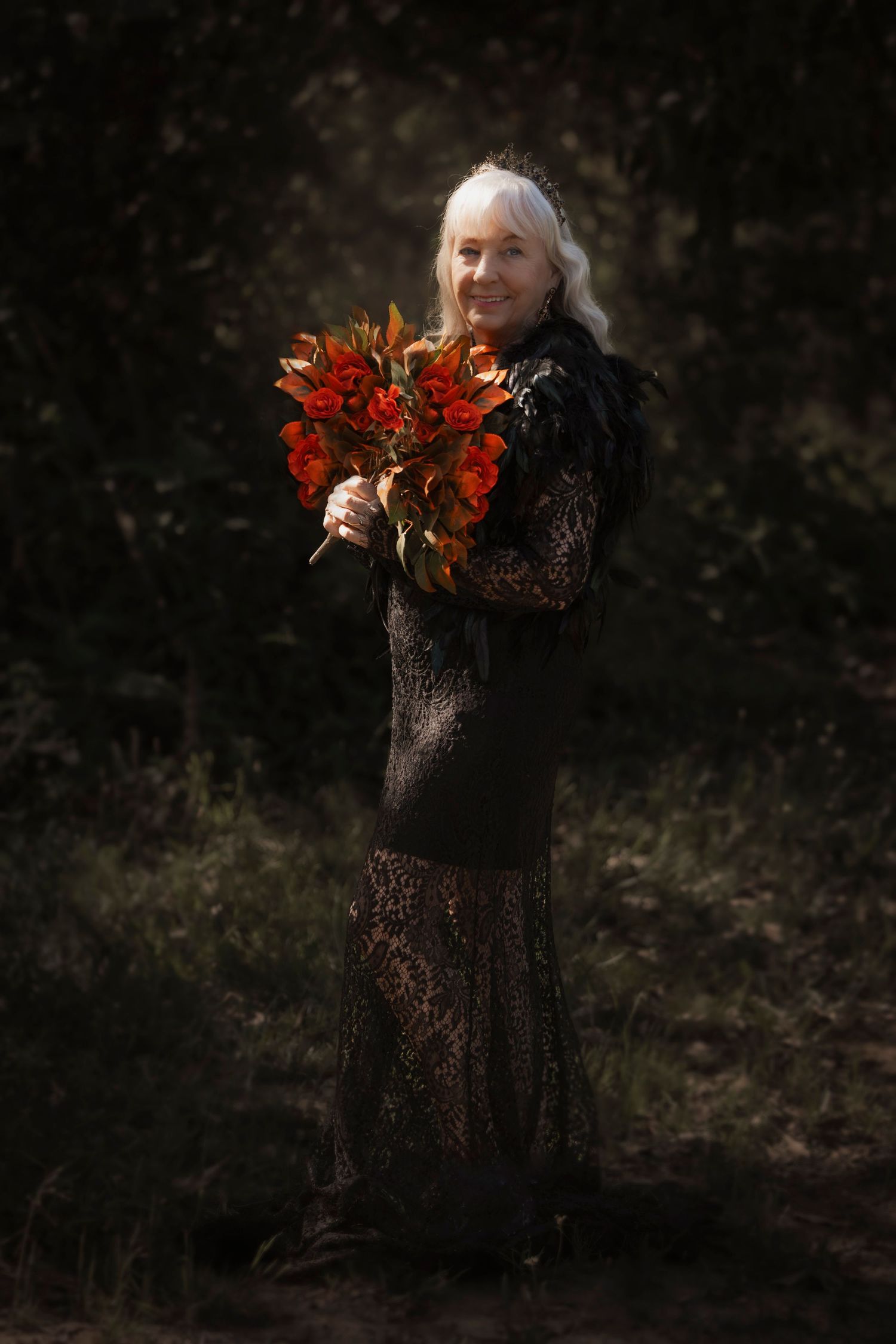 Artistic portrait in black lace dress holding vibrant red autumn flowers in dark forest setting.
