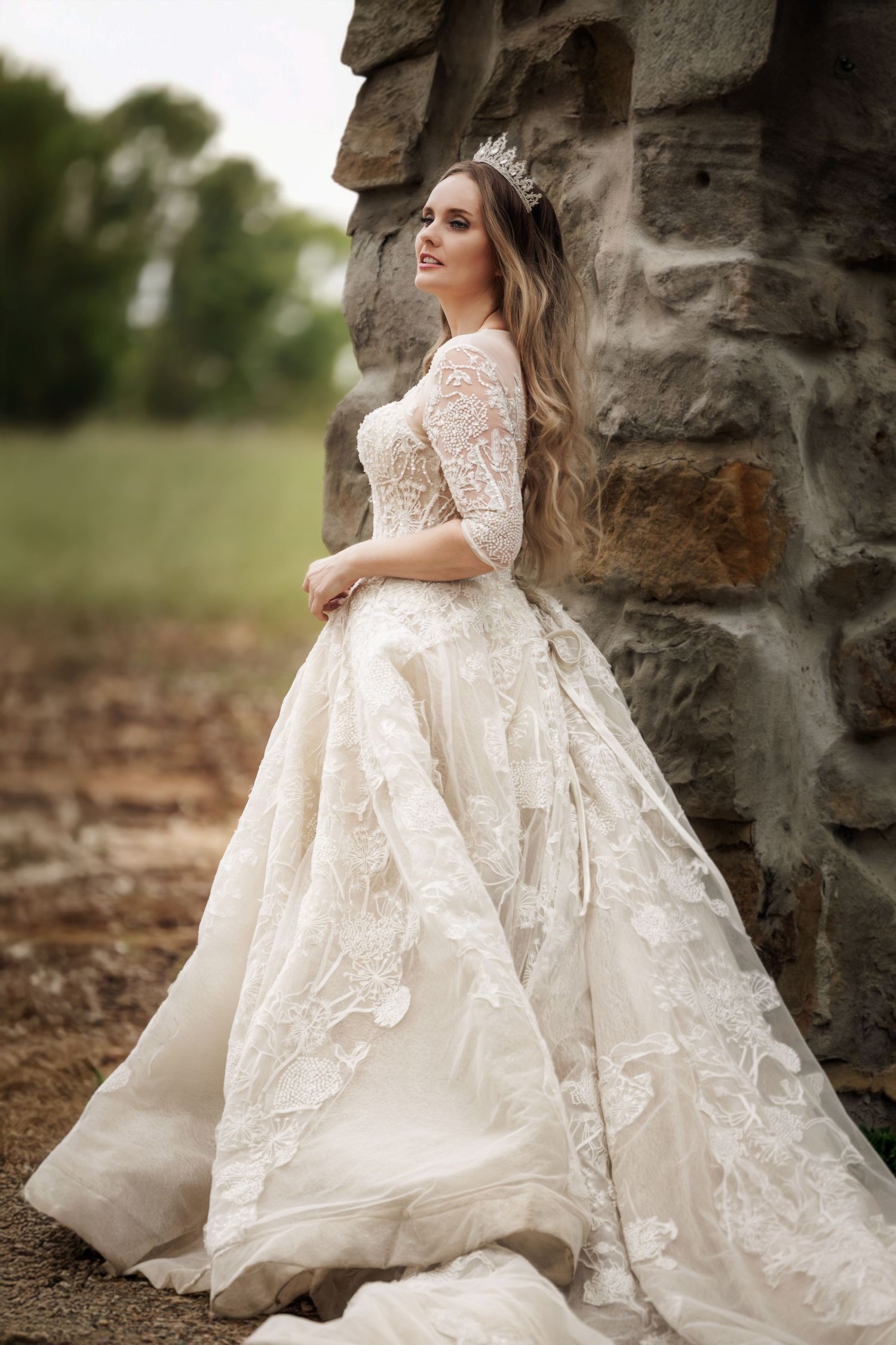 A bride in a luxurious lace wedding gown with tiara poses against a rustic stone wall in an outdoor setting.
