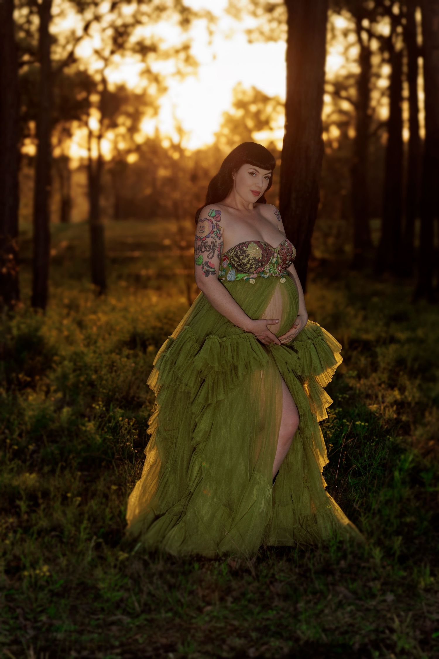 A model in a flowing green tulle gown with tattoos poses in a forest backlit by golden sunset light through pine trees.