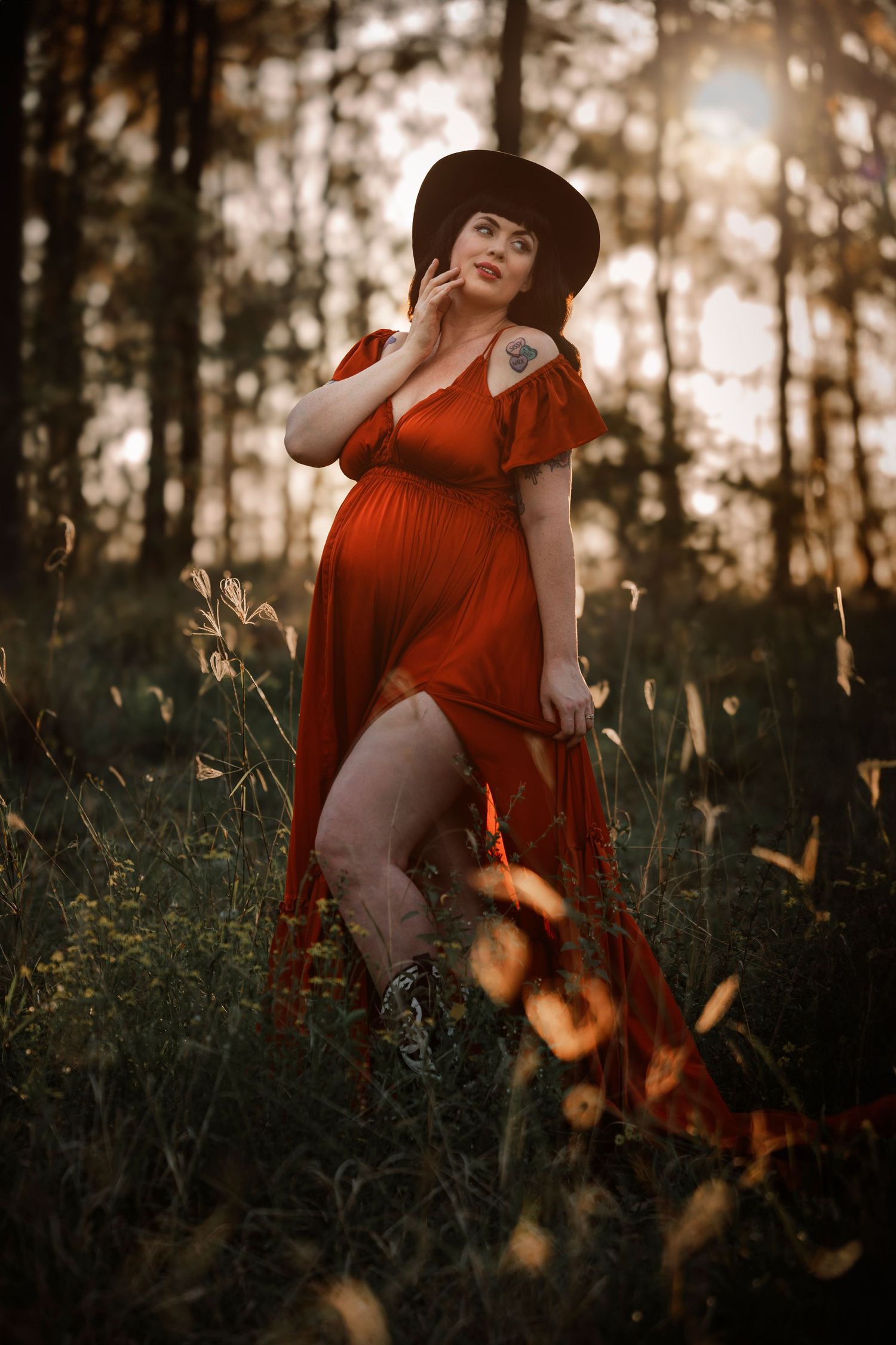 A pregnant woman in flowing rusty ochre col dress catches sunlight in a dreamy bush setting with tall trees and wild grass at golden hour.