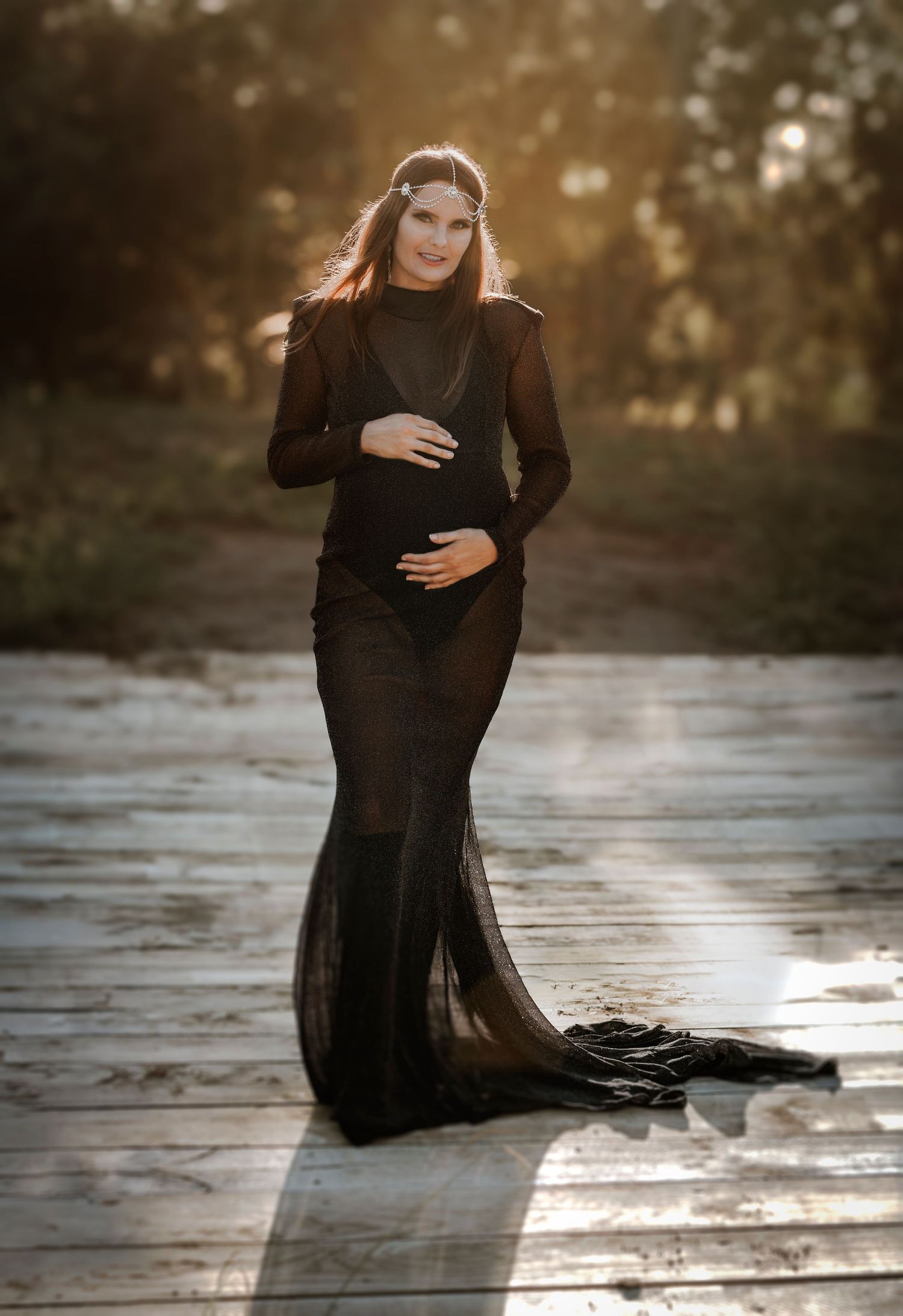 Atmospheric maternity portrait on a wooden deck at golden hour showing a woman in a flowing black dress touching her belly.