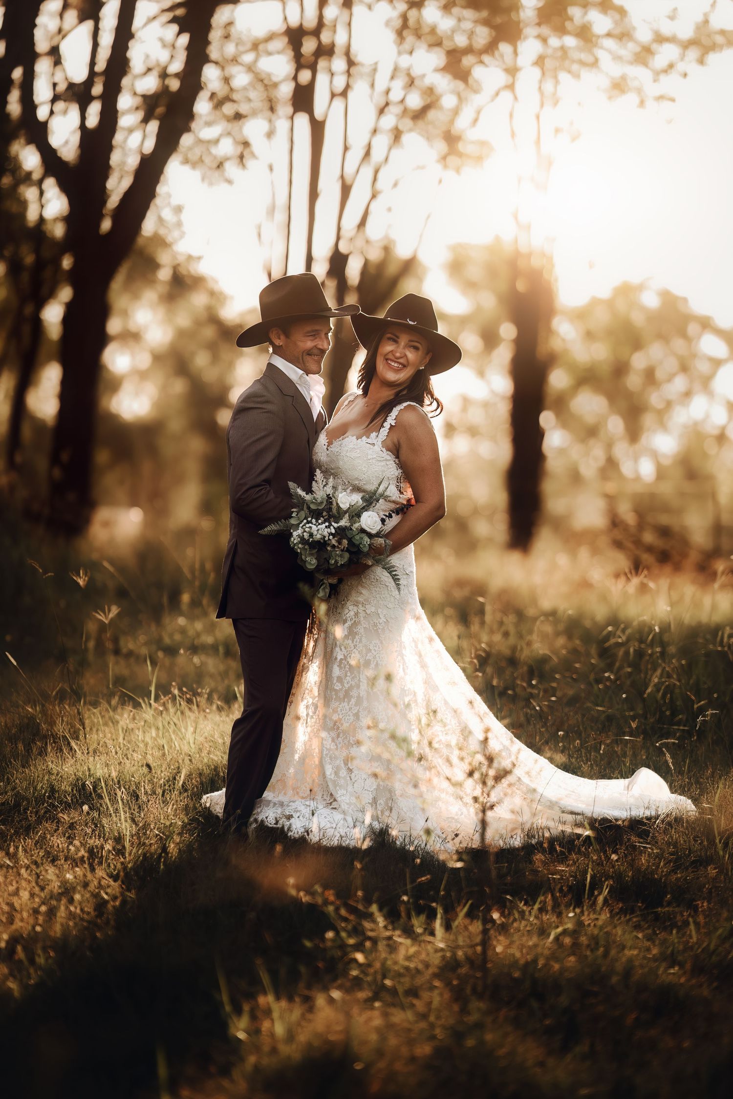 Newlywed couple shares romantic moment in sunlit forest wearing rustic wedding attire with cowboy hat and white gown.