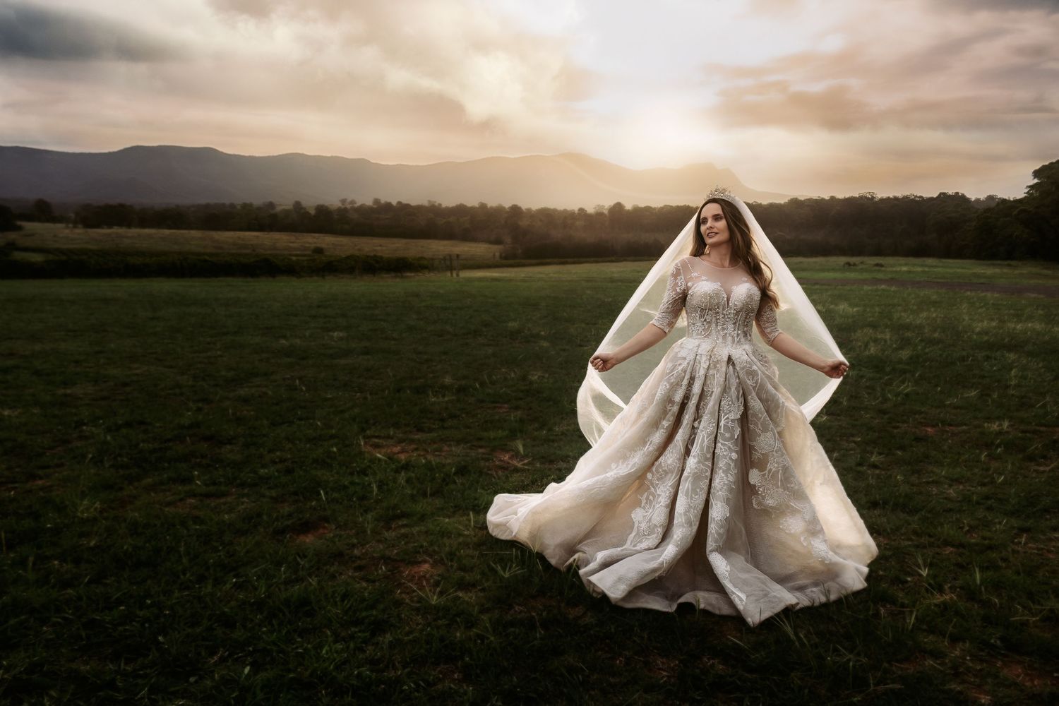 A wedding dress flows elegantly in motion against a mountain sunset backdrop in an open field.