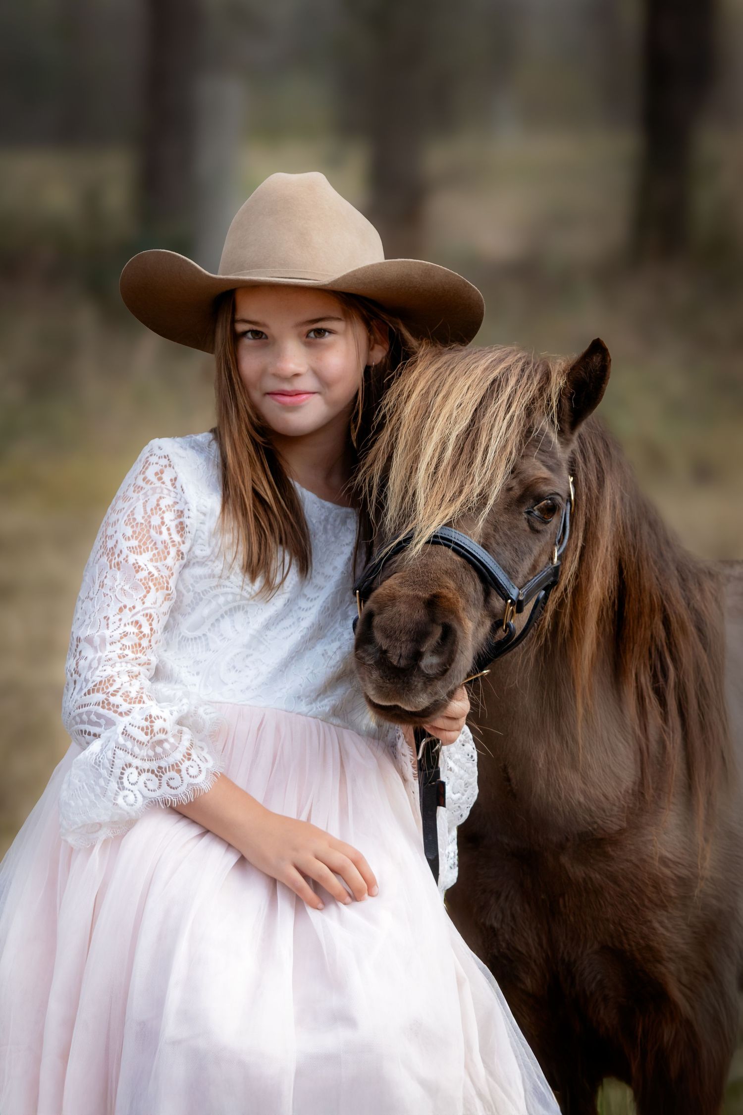 A brown pony with golden mane poses with a girl wearing a white and pink lace dress and light brown cowgirl hat in a rustic outdoor setting.