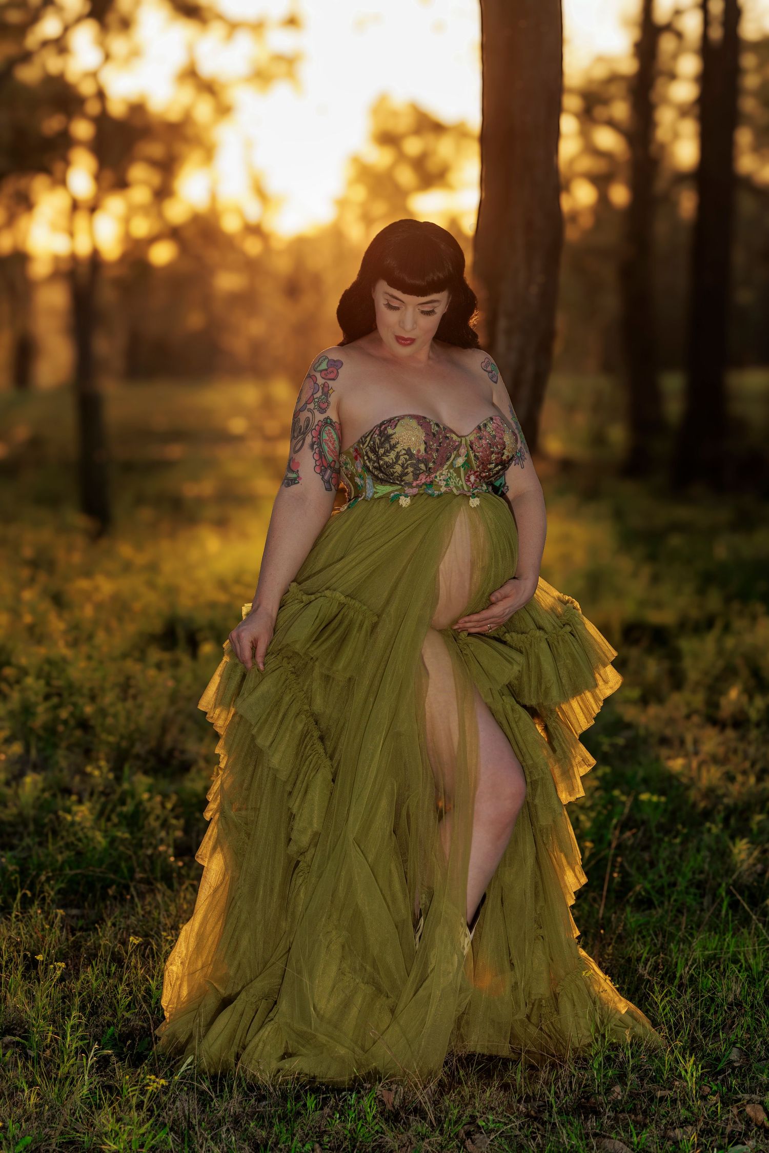A woman in a flowing olive green gown with floral details poses in a sun-dappled forest at golden hour.