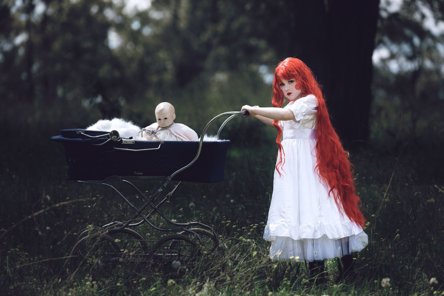 A figure in a white dress with vibrant red hair stands in a moody outdoor setting with dark trees and fog.