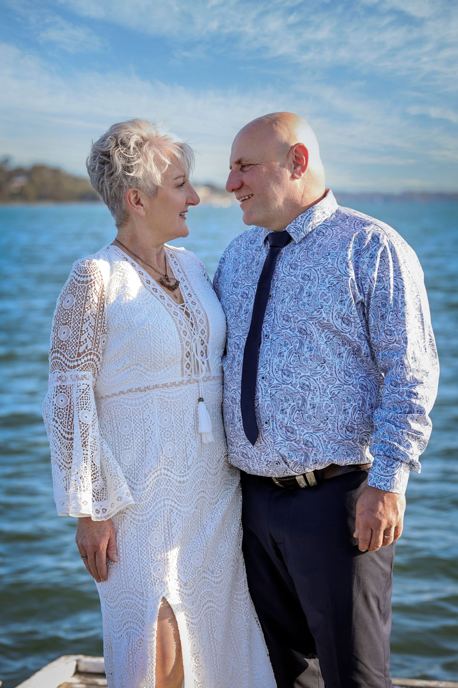 A couple stands together by the water, dressed in white lace and blue patterned attire on a sunny day.