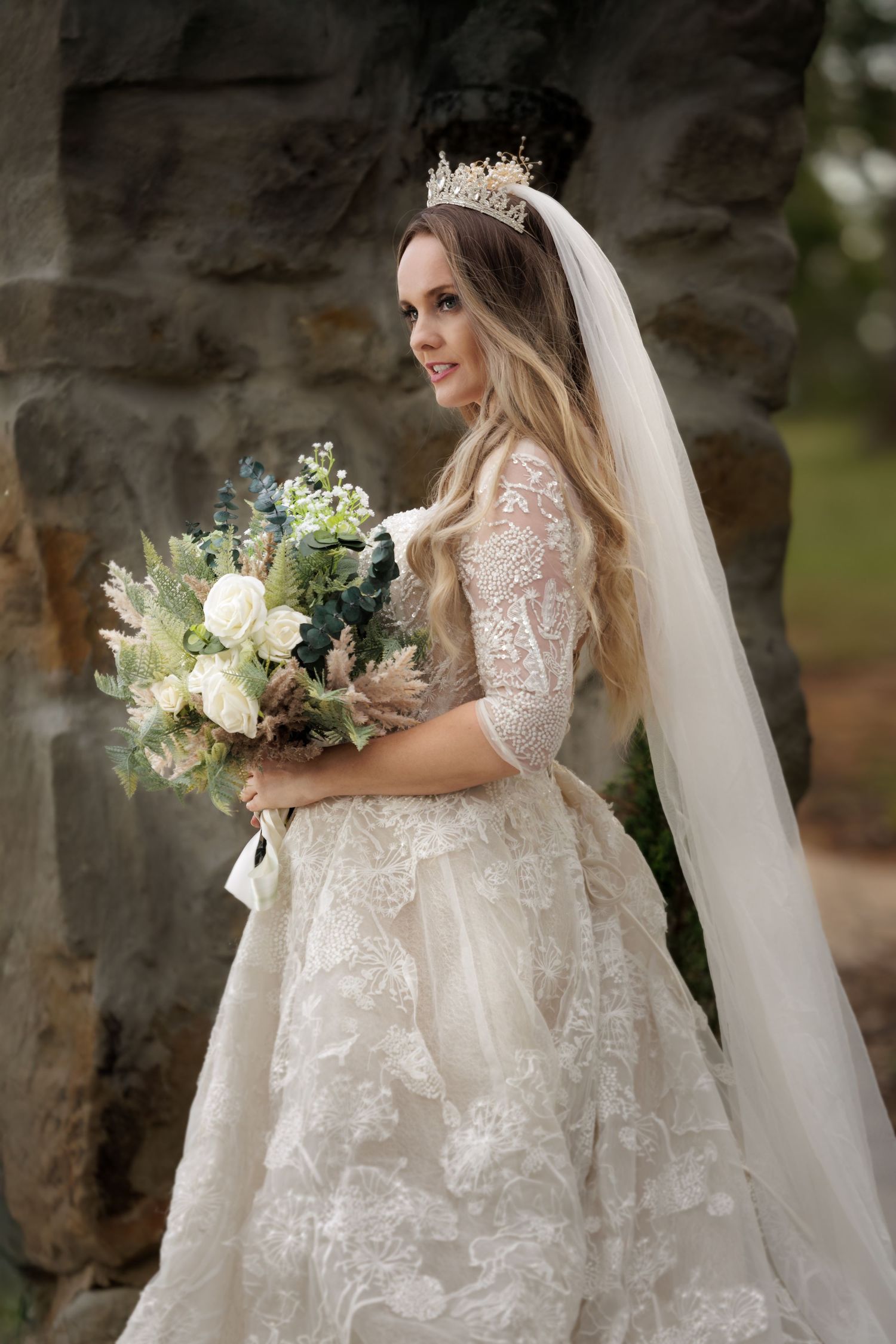 A bride in a lace wedding gown with a tiara holds a white and green floral bouquet against a stone wall backdrop.