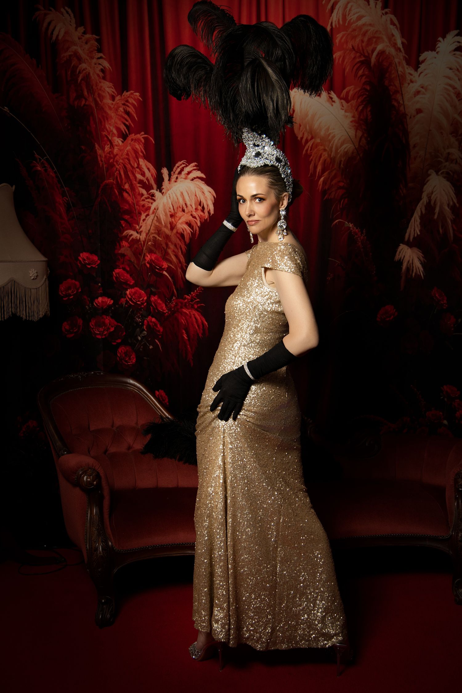 A performer in a sparkly gold gown poses dramatically against a dark red backdrop with feather decorations.