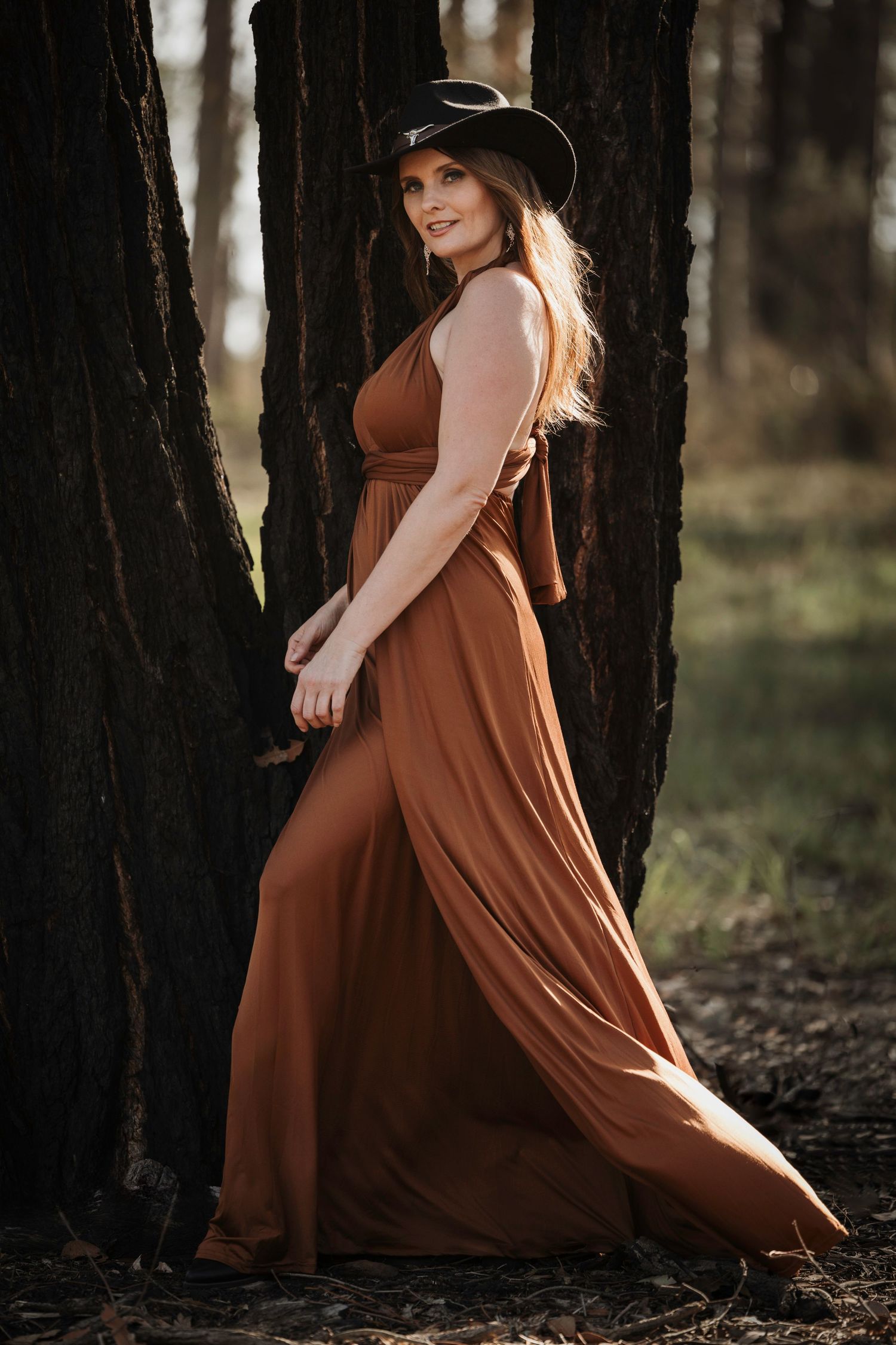 Rustic western-inspired photoshoot featuring flowing rust-colored maxi dress and cowboy hat among trees.