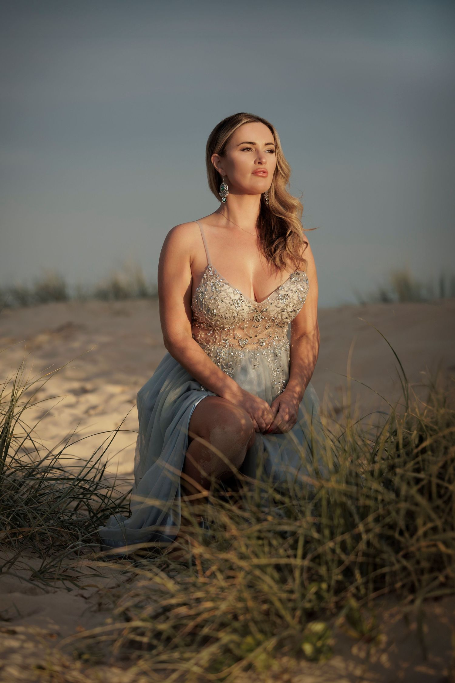 A person in an elegant blue evening gown poses at sunset among sand dunes and beach grass.