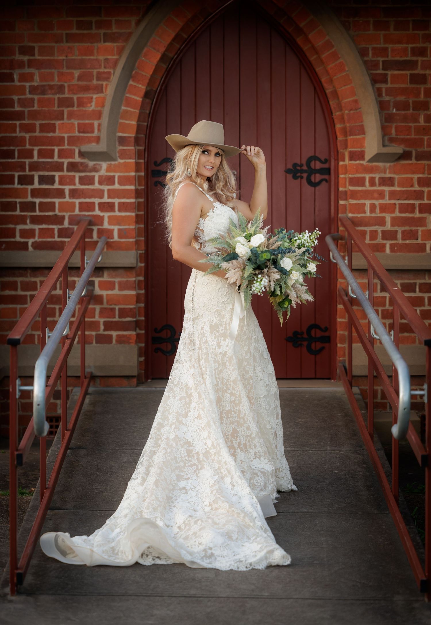A rustic bride in a lace wedding gown and cowboy hat poses with a white floral bouquet against an arched church door.